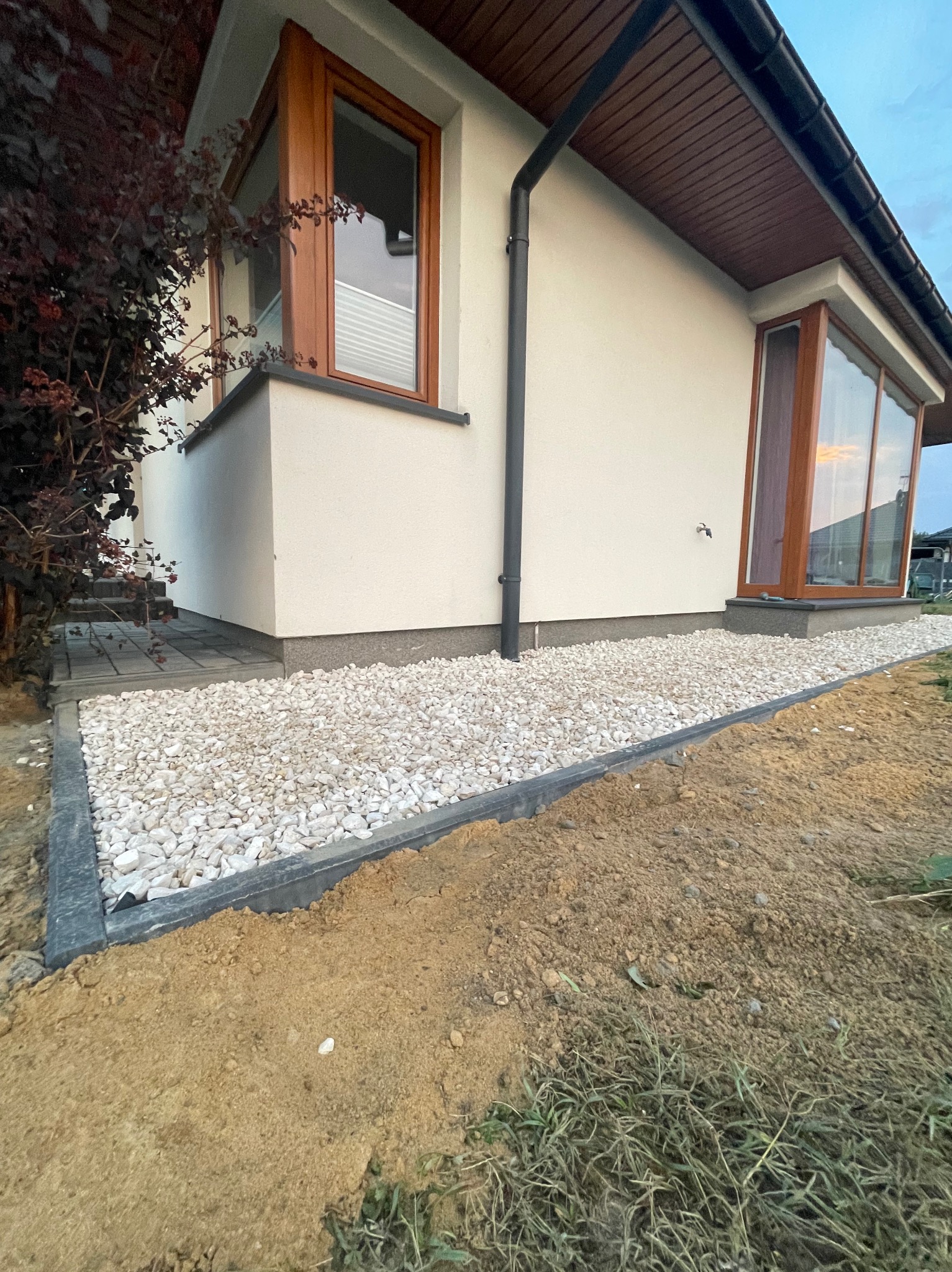 Wykończona ścieżka z białego kamienia przy domu z brązowymi oknami, obrzeża z tworzywa. Widoczna ziemia i trawa. Ujęcie z dołu.