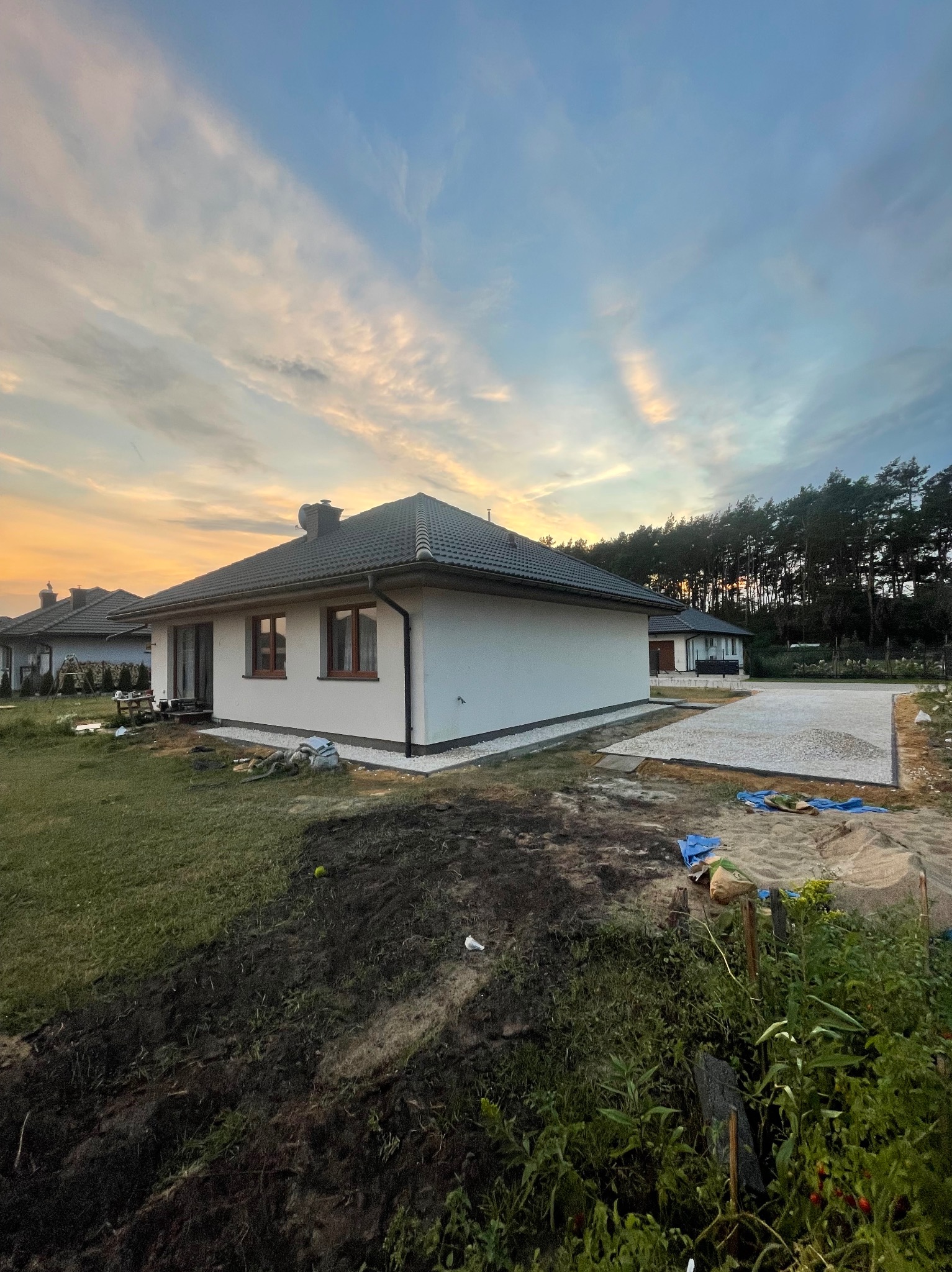 Nowoczesny dom jednorodzinny z przygotowanym podłożem pod taras, widok z boku na tle zachodzącego słońca i lasu. Przygotowanie terenu pod budowę.