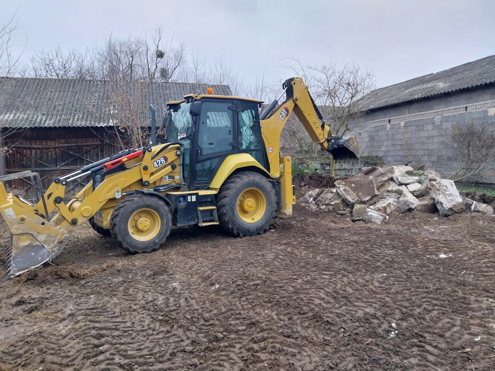 Żółta koparko-ładowarka CAT 428 przy pryzmie gruzu na błotnistym terenie, widoczne ślady opon, w tle budynki gospodarcze pod szarym niebem.
