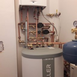 Drobne usługi hydrauliczne - Biały zasobnik ciepłej wody użytkowej Keller z miedzianą instalacją, widoczne pompy obiegowe i zawory. Na ścianie wiszący kocioł grzewczy.