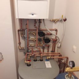 Drobne usługi hydrauliczne - Kocioł grzewczy z rozbudowaną instalacją hydrauliczną z miedzianych rur, zaworów i pomp. Widoczne elementy kotła, zbiornik i osprzęt. Białe ściany w tle.