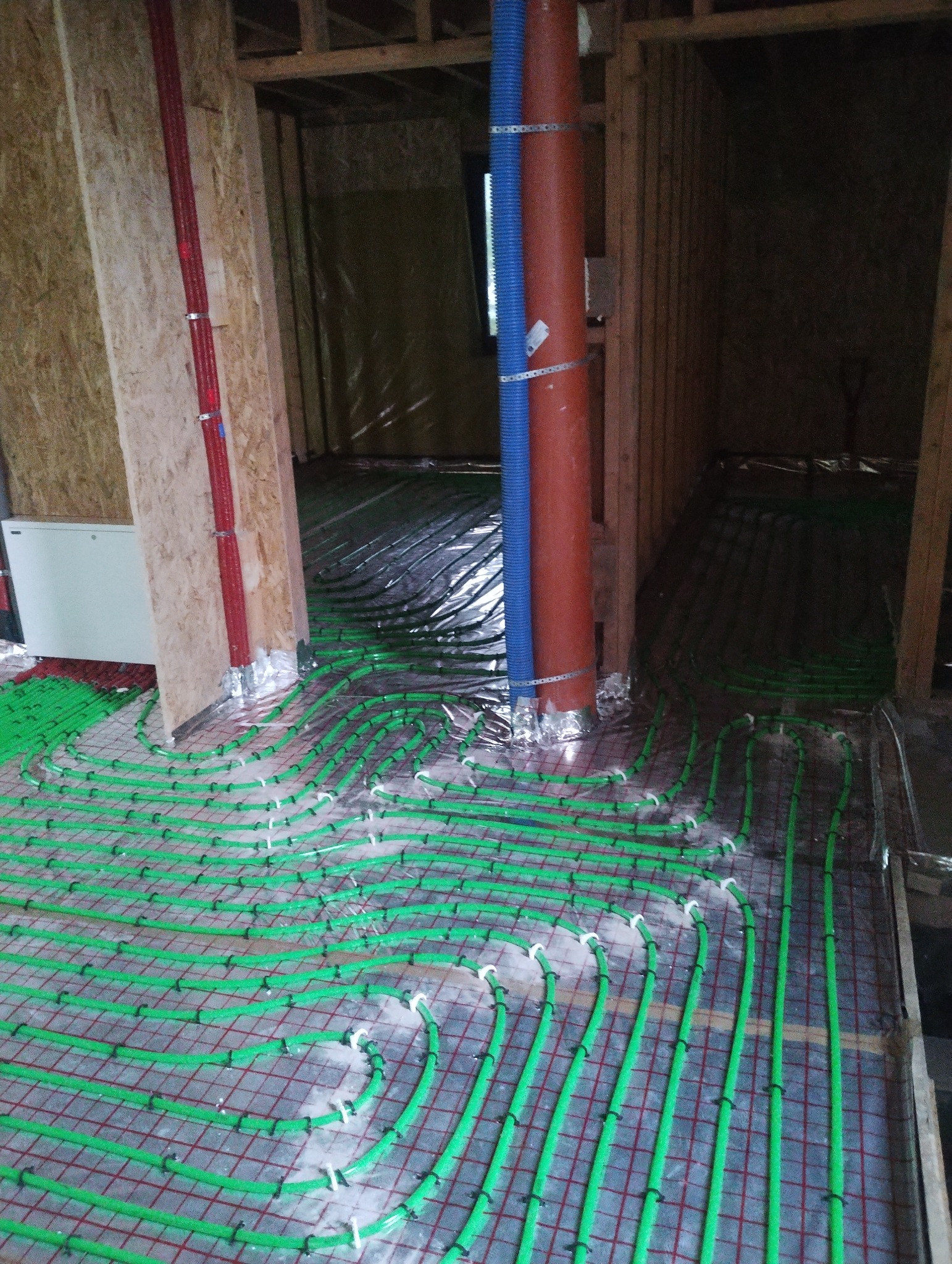 Zielone rury ogrzewania podłogowego ułożone na folii izolacyjnej, widoczne elementy konstrukcji drewnianej i pionowe rury instalacyjne w kolorach czerwonym i niebieskim.