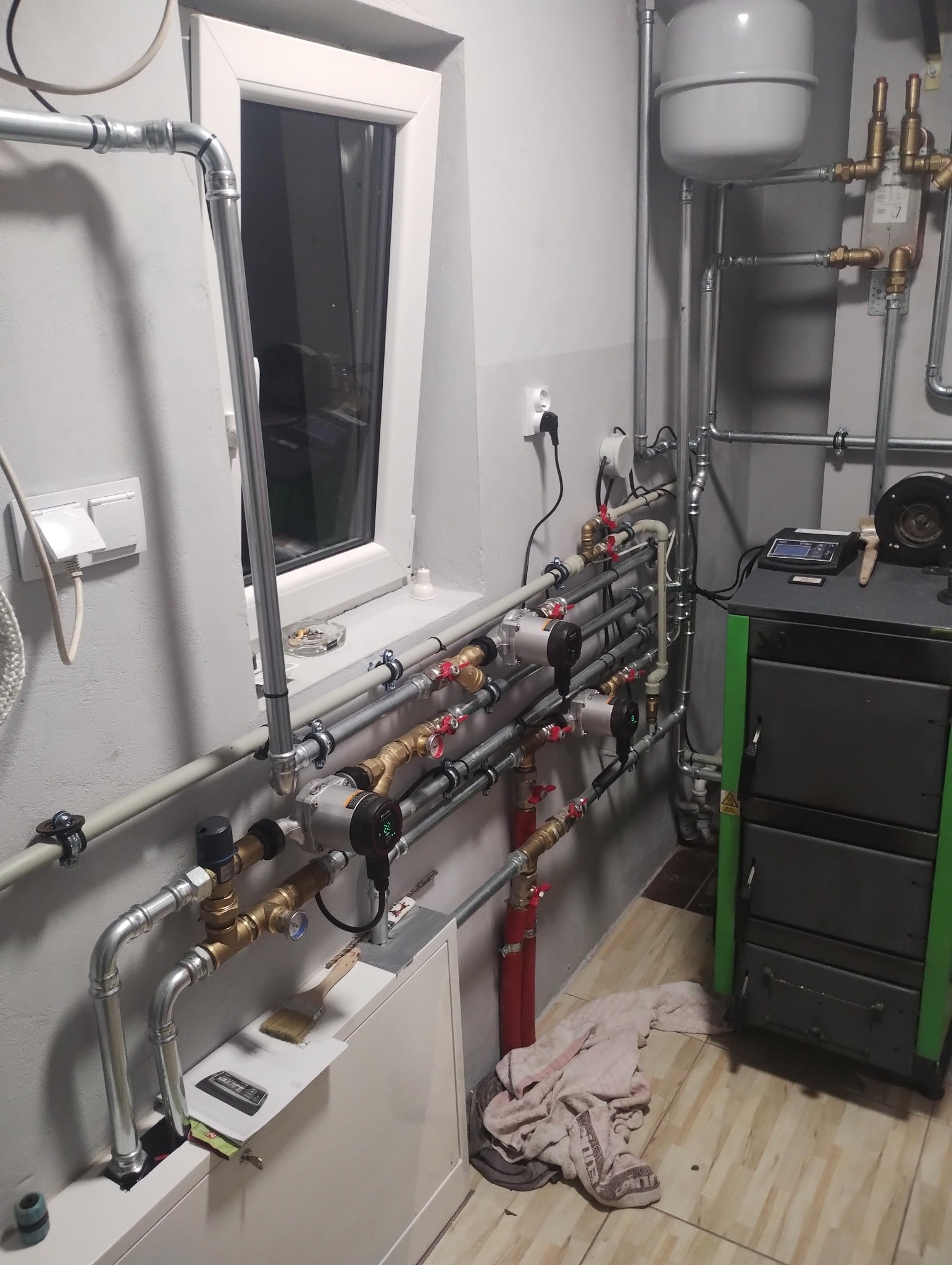 Złożona instalacja grzewcza z kotłem na paliwo stałe, rurami miedzianymi i stalowymi, pompami obiegowymi, zbiornikiem wyrównawczym w kotłowni z oknem.