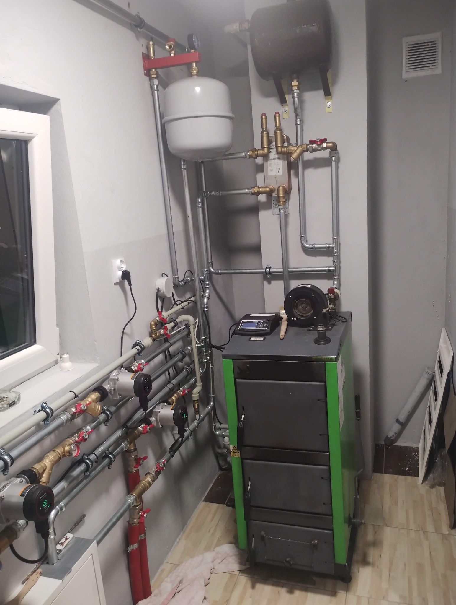 Kotłownia z piecem na paliwo stałe, zbiornikami wyrównawczymi i rozbudowaną instalacją grzewczą z licznymi zaworami i pompami obiegowymi. Szare ściany, okno w tle.