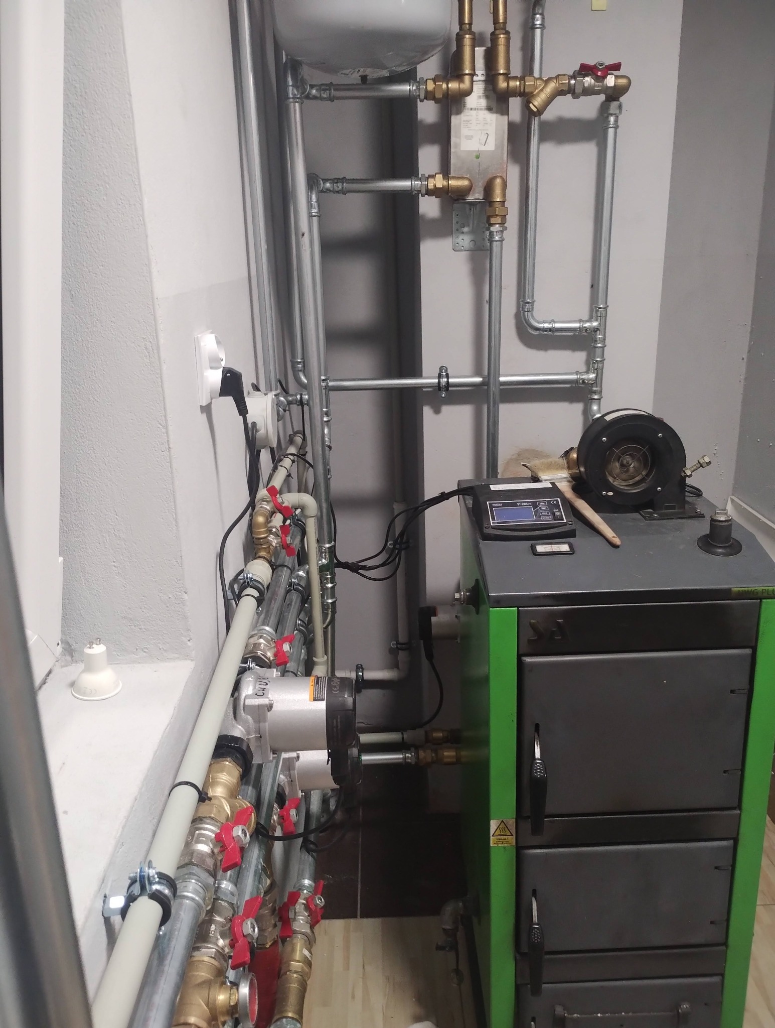 Złożona instalacja grzewcza z kotłem na paliwo stałe, rurami miedzianymi i stalowymi, zaworami z czerwonymi pokrętłami oraz pompą obiegową. Widoczne elementy sterujące kotłem.