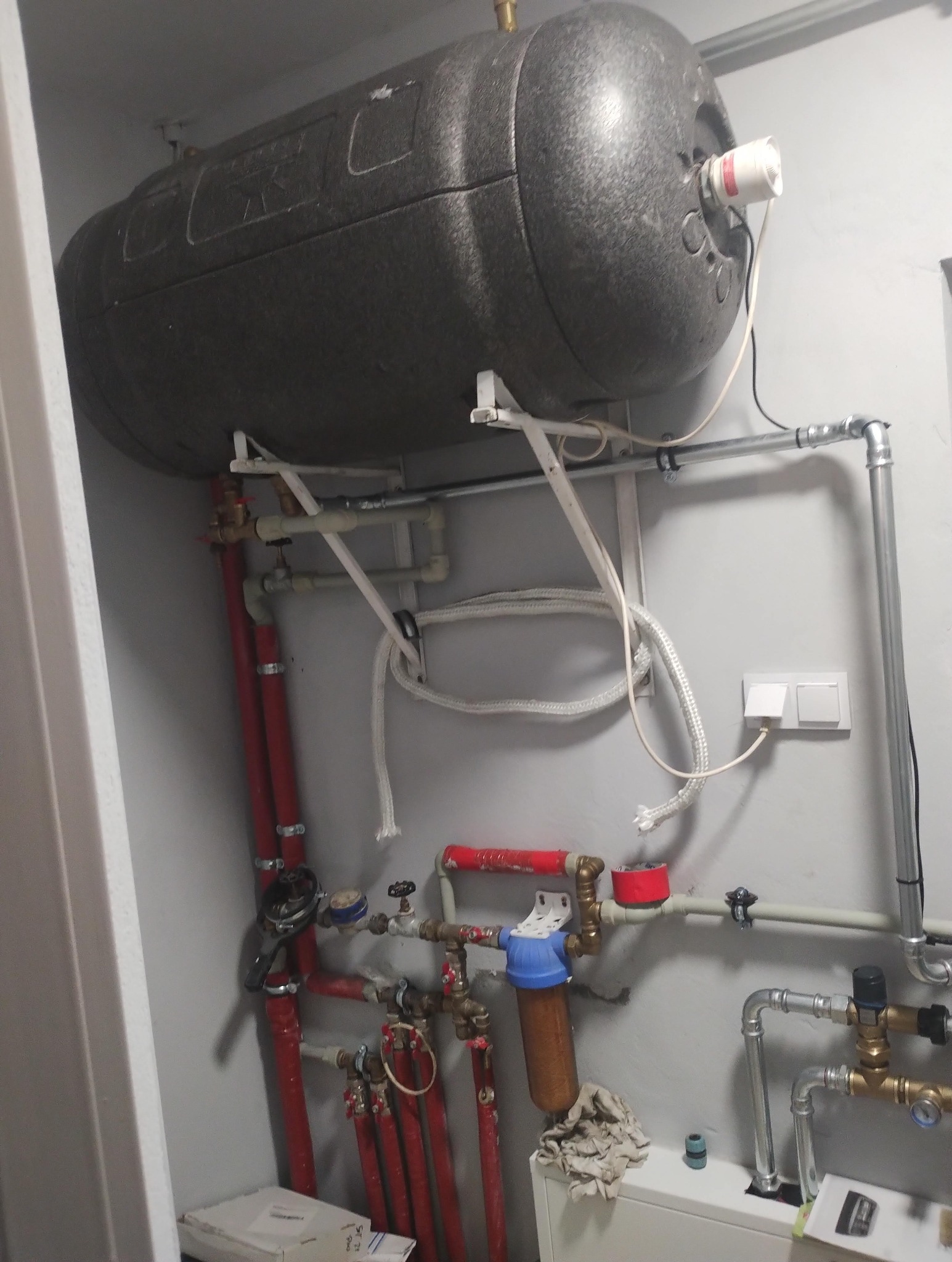Szary, poziomy zbiornik na wodę, zamontowany pod sufitem, z licznymi rurami (czerwone, srebrne, białe) i zaworami. Widoczny filtr wody i fragment ściany pomalowanej na szaro.