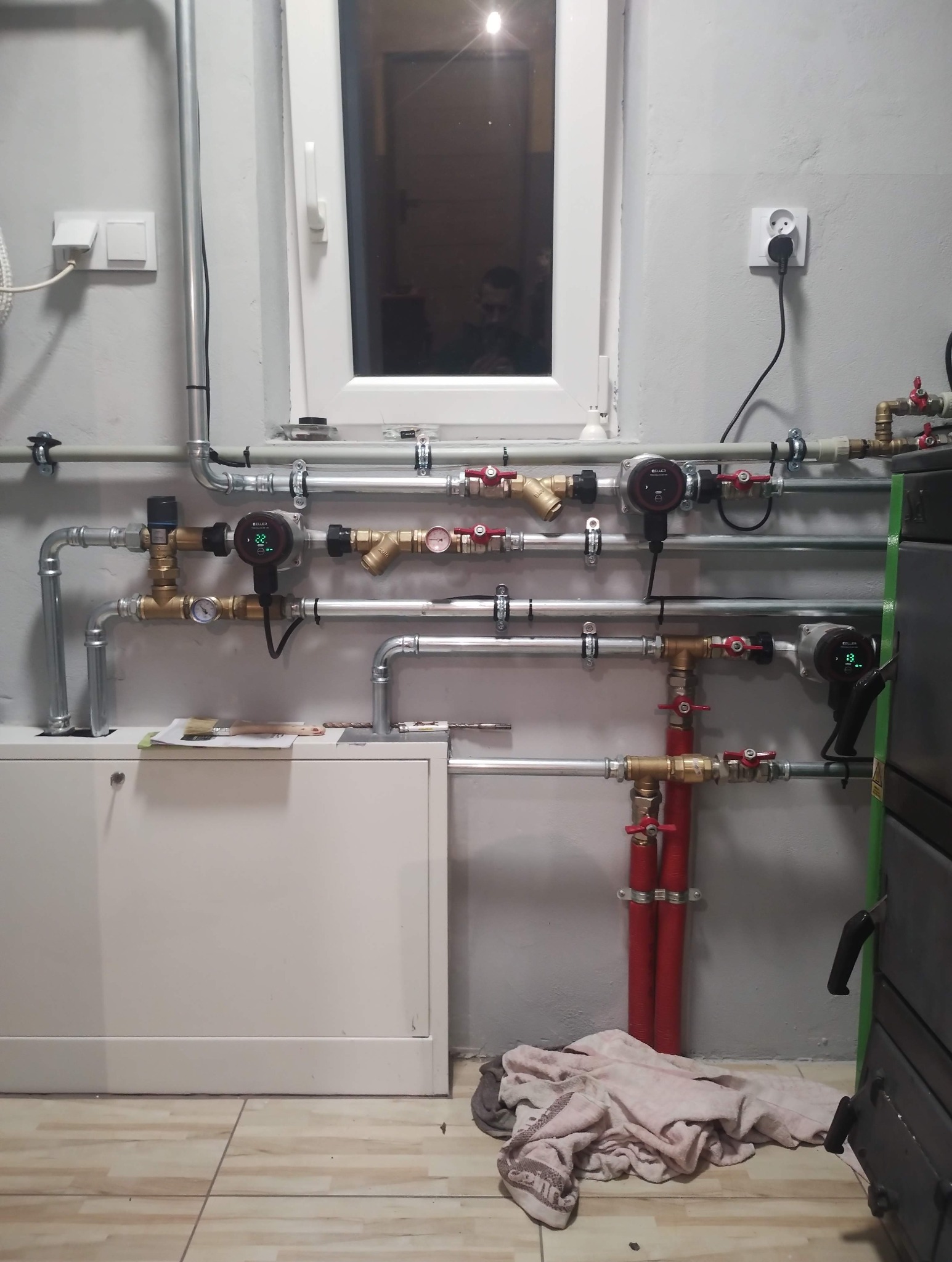 Złożona instalacja grzewcza z licznymi rurami, zaworami i pompami obiegowymi na szarej ścianie, widoczna w kotłowni. W tle okno z odbiciem osoby.