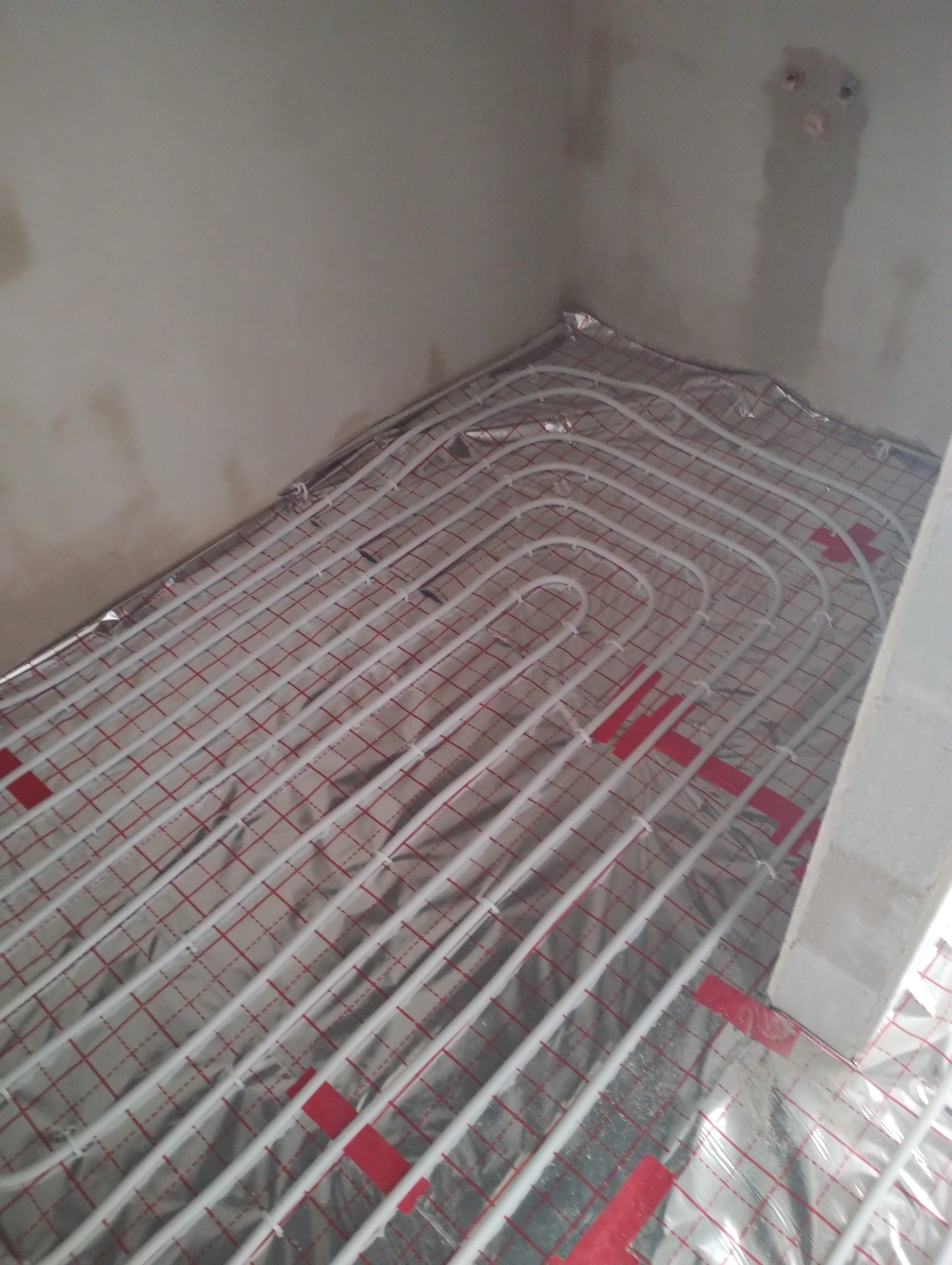 Białe rury ogrzewania podłogowego ułożone na srebrnej folii z czerwoną siatką, przymocowane czerwonymi taśmami, w surowym wnętrzu przed wylewką.