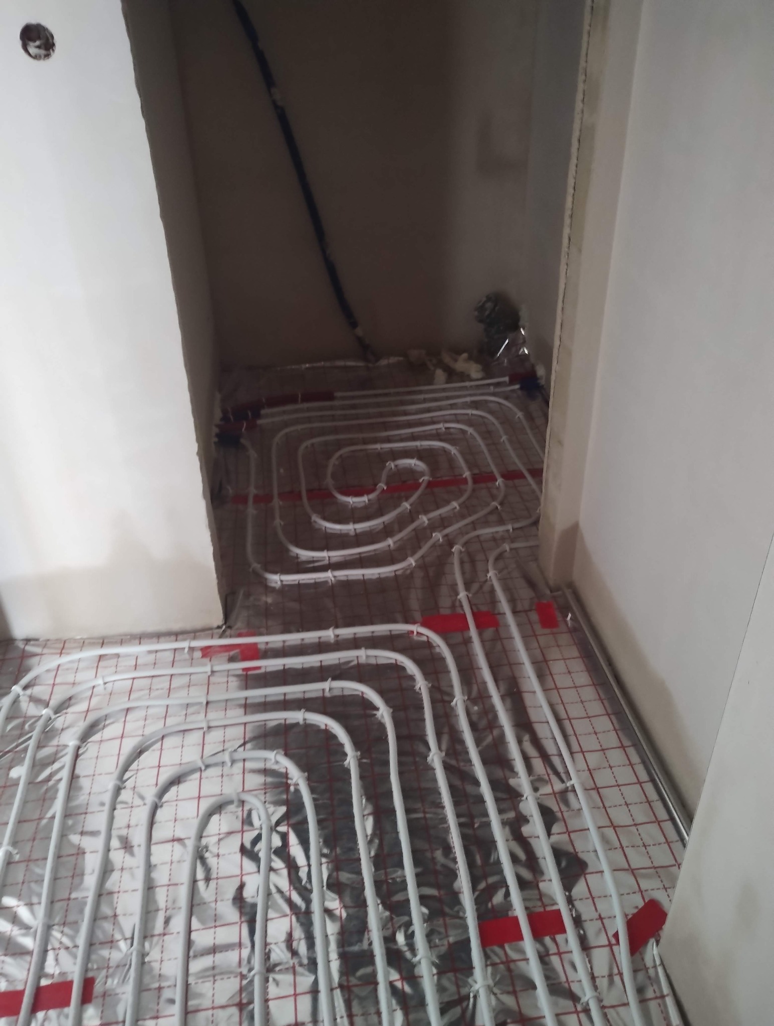 Białe rury ogrzewania podłogowego ułożone spiralnie na srebrnej folii z czerwoną siatką w surowym wnętrzu. Widoczne fragmenty ścian i instalacji.