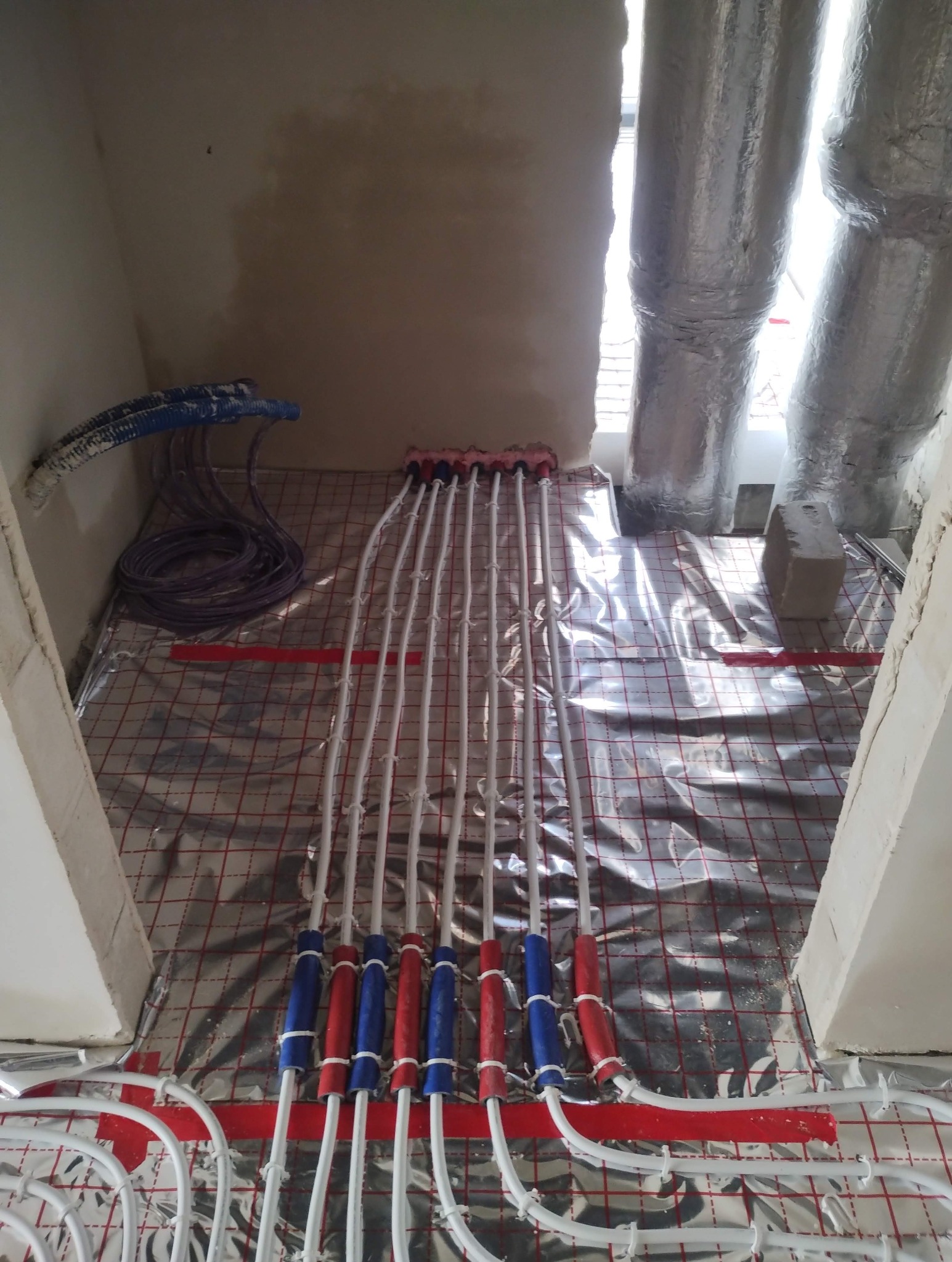 Białe rury ogrzewania podłogowego, z czerwonymi i niebieskimi oznaczeniami, ułożone na srebrnej folii z czerwoną siatką, w surowym wnętrzu przed wylewką.
