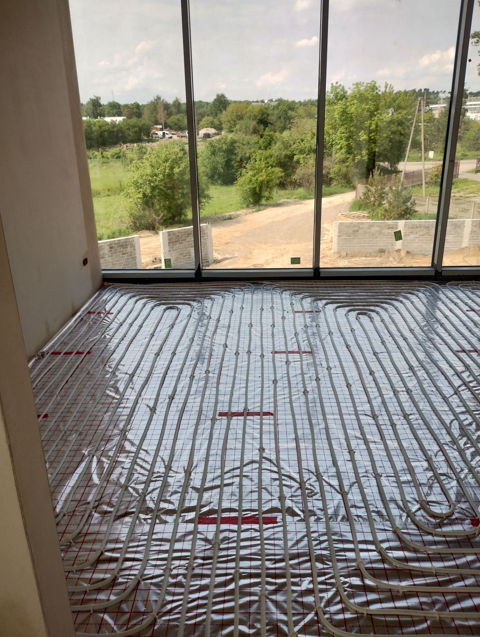 Ułożona instalacja ogrzewania podłogowego na folii aluminiowej w surowym wnętrzu z widokiem na zieleń za oknem. Widoczne pętle rur i czerwone elementy mocujące.
