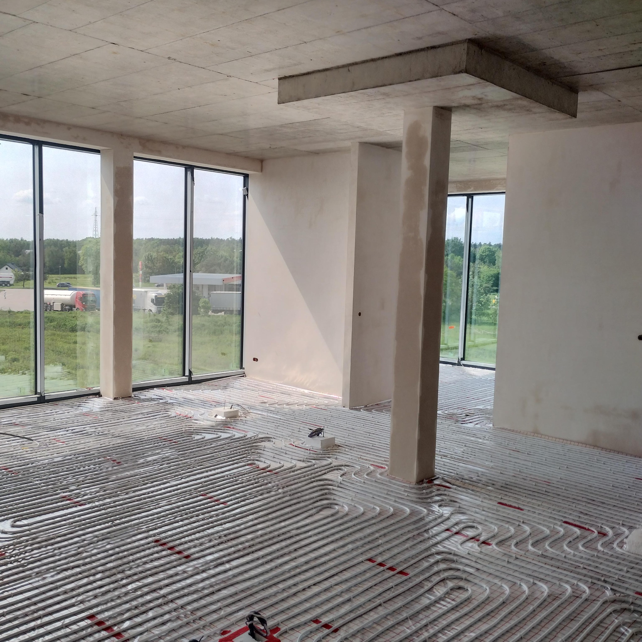 Rozłożona instalacja ogrzewania podłogowego w surowym wnętrzu z dużymi oknami. Widoczne rury grzewcze na podkładzie izolacyjnym, betonowy strop i kolumny.