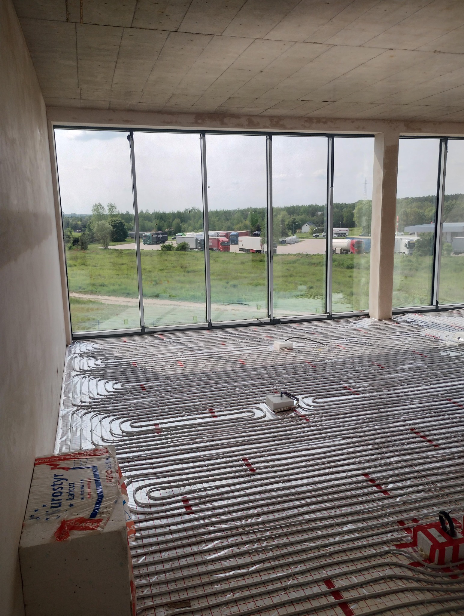 Instalacja ogrzewania podłogowego w Lublinie, widok na ułożone rury na folii izolacyjnej w surowym wnętrzu z dużym oknem i widokiem na zieleń.