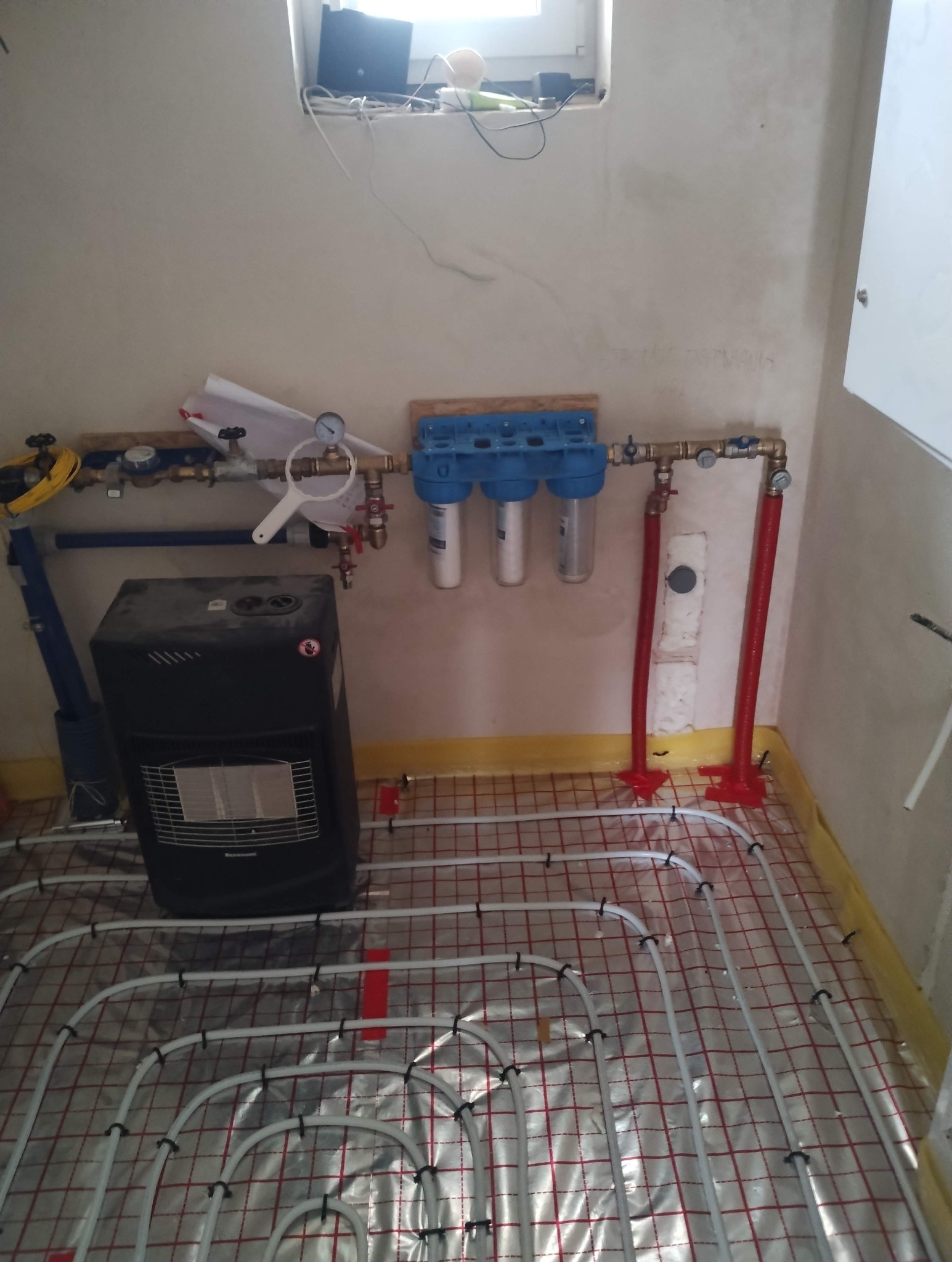 Instalacja ogrzewania podłogowego w trakcie montażu, widoczne rury na folii aluminiowej, filtry wody i grzejnik przenośny. Prace hydrauliczne w pomieszczeniu.