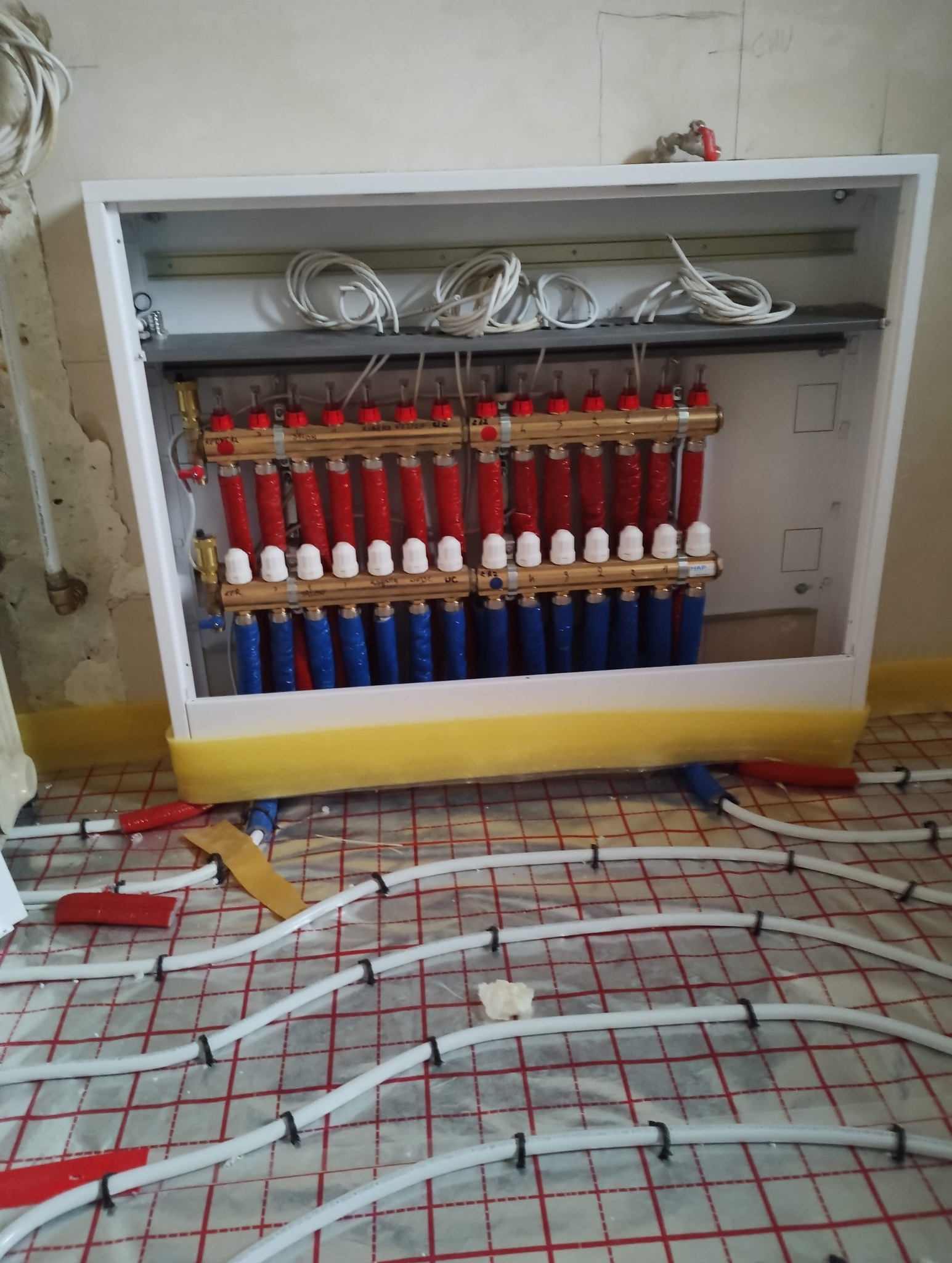 Rozdzielacz ogrzewania podłogowego w białej szafce, rury ułożone na folii z rastrem. Widoczne czerwone i niebieskie elementy instalacji. Żółta taśma zabezpieczająca.
