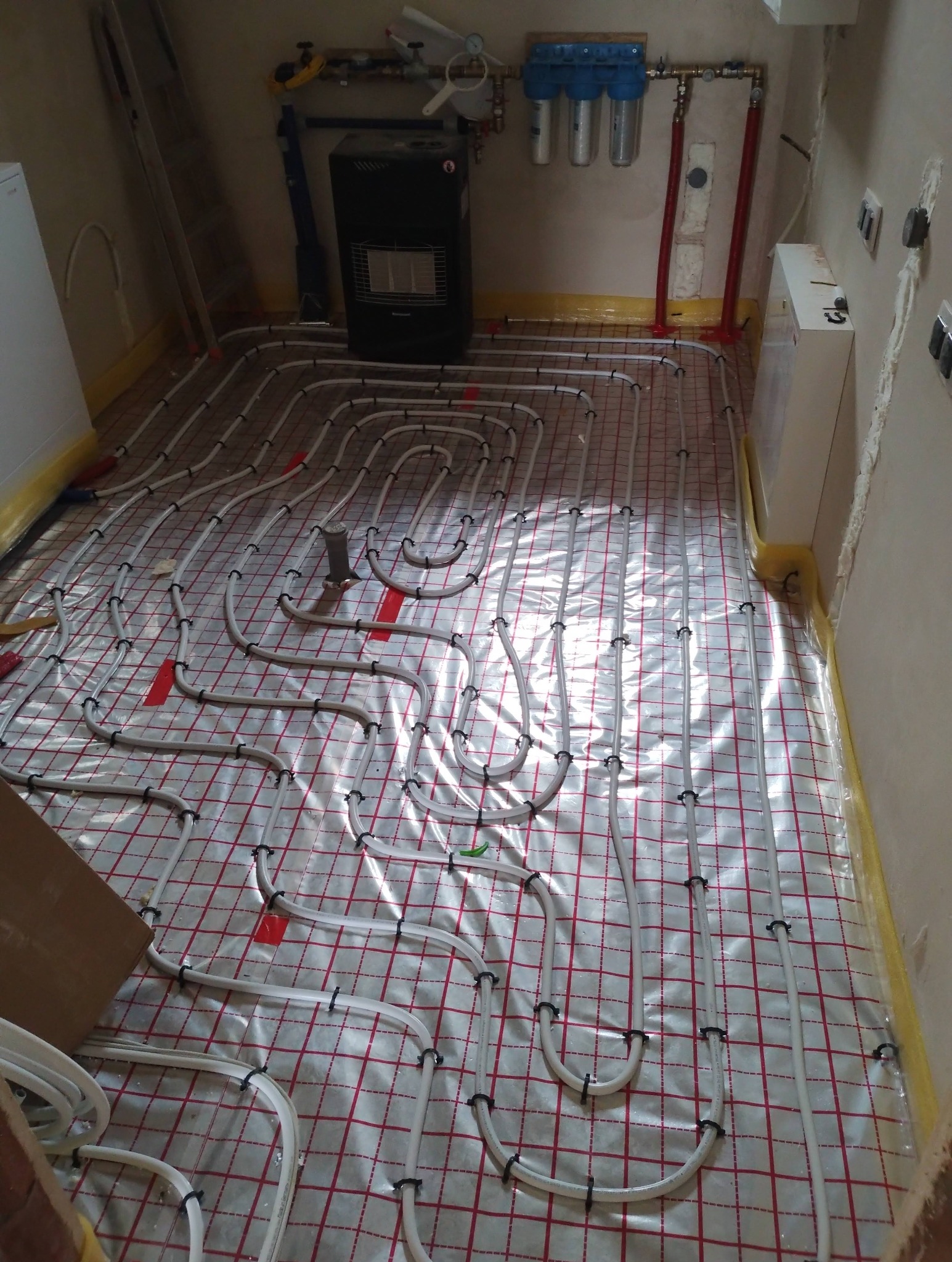 Białe rury ogrzewania podłogowego ułożone na folii z czerwoną kratką, widoczne elementy instalacji grzewczej i filtry wody. Ujęcie z góry, w pomieszczeniu w trakcie prac.