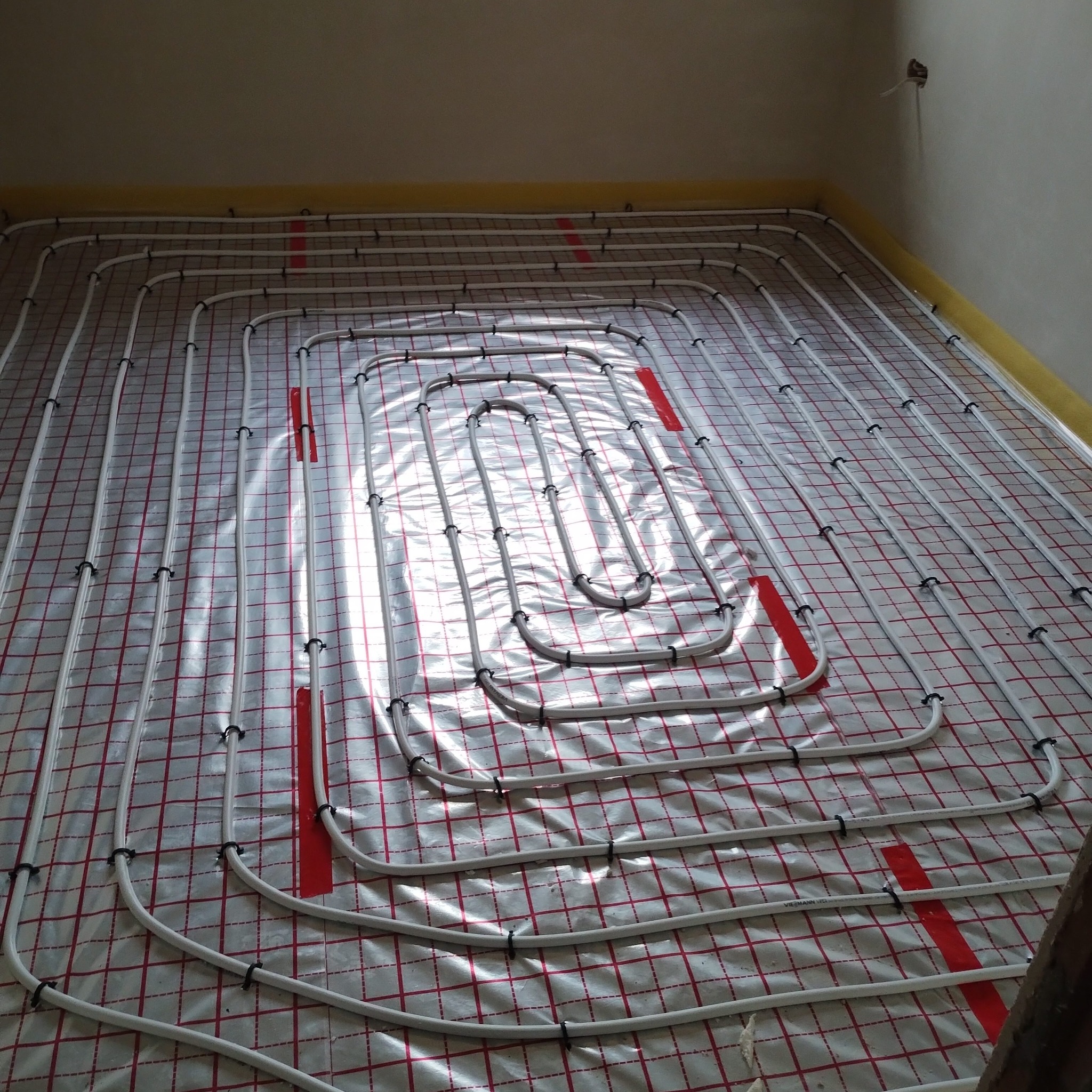 Białe rury ogrzewania podłogowego ułożone na folii z czerwoną kratką, przymocowane czarnymi klipsami. Widok z góry na instalację przed wylaniem posadzki.