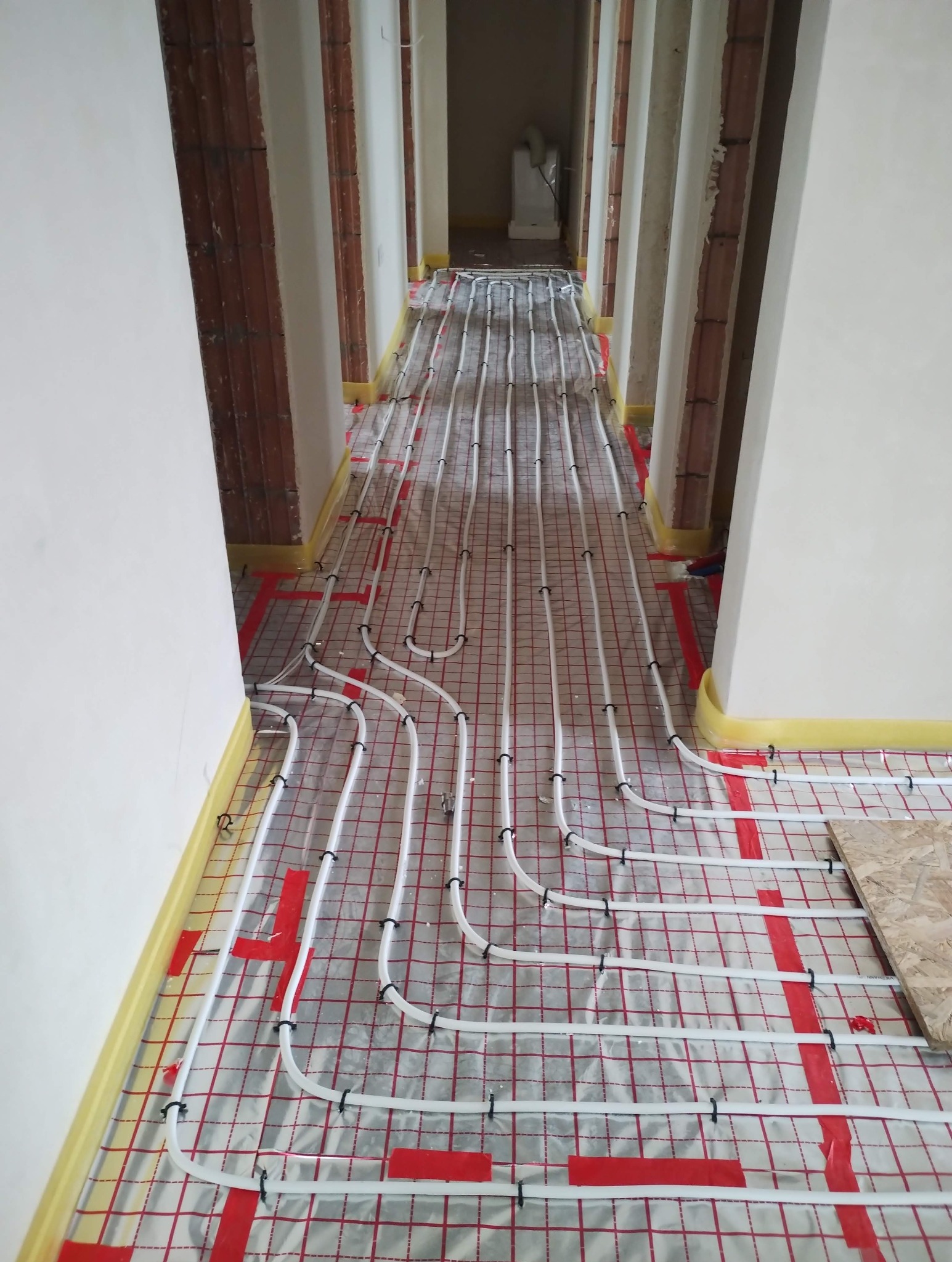 Białe rury ogrzewania podłogowego, zamocowane na czerwonej siatce, rozłożone w korytarzu w trakcie remontu. Widoczne fragmenty ścian i przygotowane otwory na drzwi.
