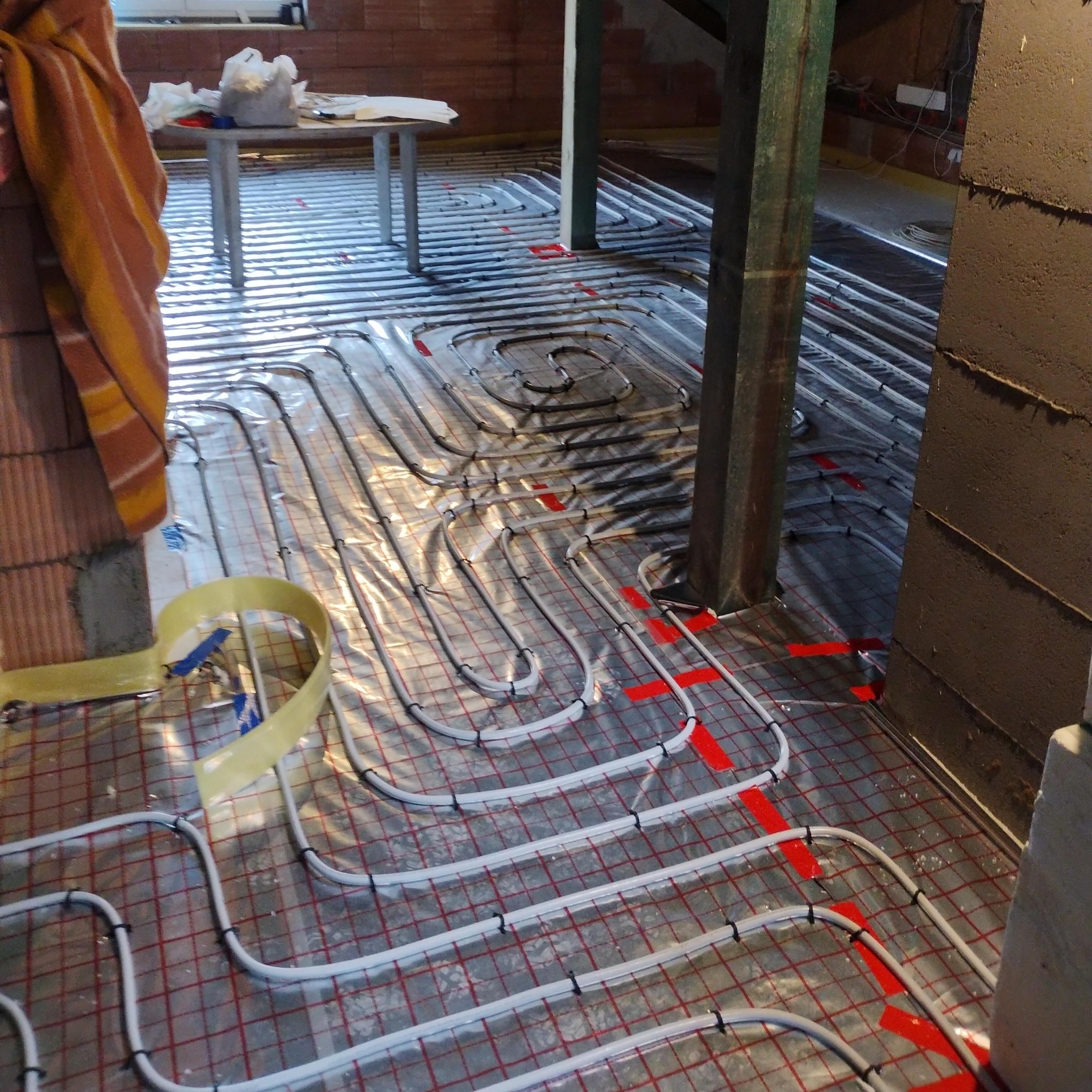 Ułożone rury ogrzewania podłogowego na folii z czerwoną siatką, przymocowane czarnymi klipsami. Widoczne fragmenty ściany z cegły i drewniany słup.