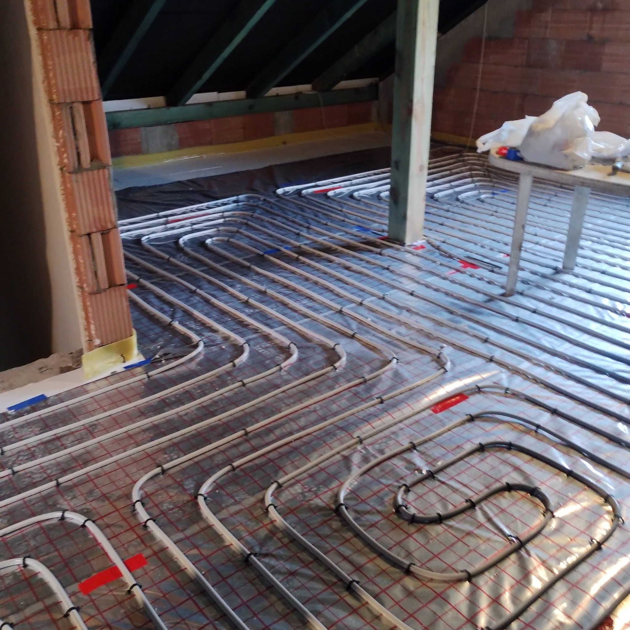 Instalacja ogrzewania podłogowego w trakcie montażu na folii izolacyjnej z widocznymi rurami grzewczymi, mocowaniami i elementami konstrukcyjnymi poddasza.