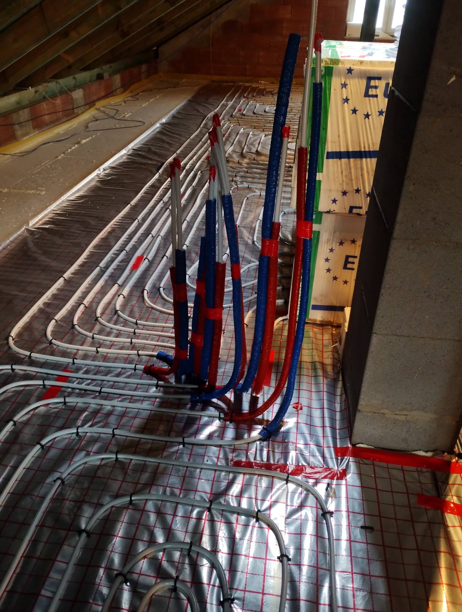 Rozłożona instalacja ogrzewania podłogowego na folii z siatką, z pionowymi rurami w izolacji czerwono-niebieskiej, widok z góry, poddasze.