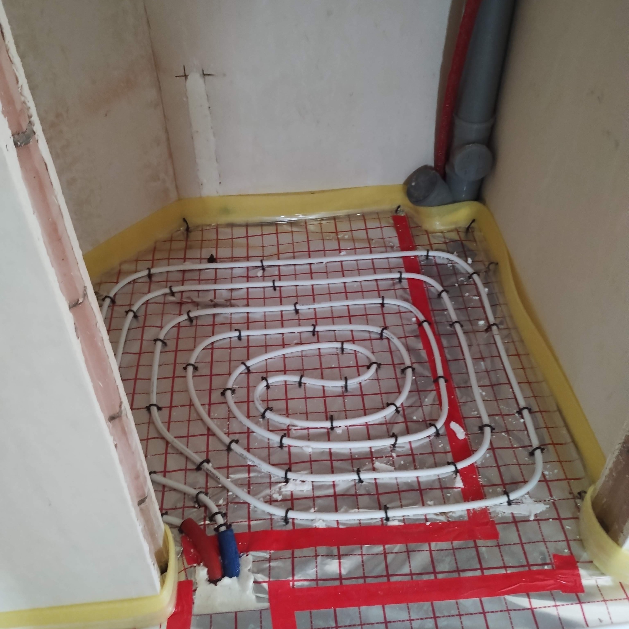 Białe rury ogrzewania podłogowego ułożone spiralnie na folii z czerwoną siatką, przymocowane czarnymi uchwytami. Widoczne fragmenty ścian i rur kanalizacyjnych.