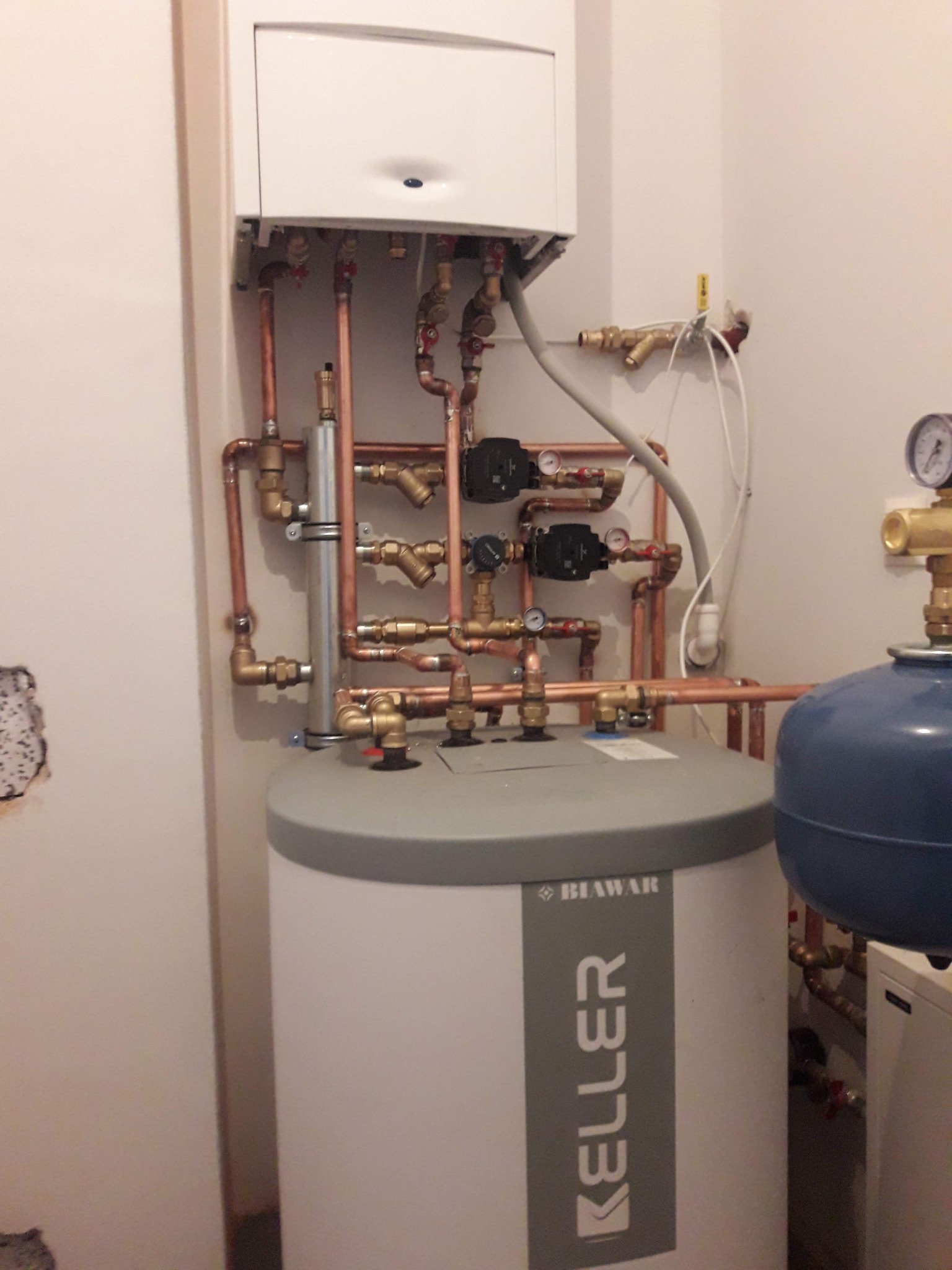 Biały zasobnik ciepłej wody użytkowej Keller z miedzianą instalacją, widoczne pompy obiegowe i zawory. Na ścianie wiszący kocioł grzewczy.