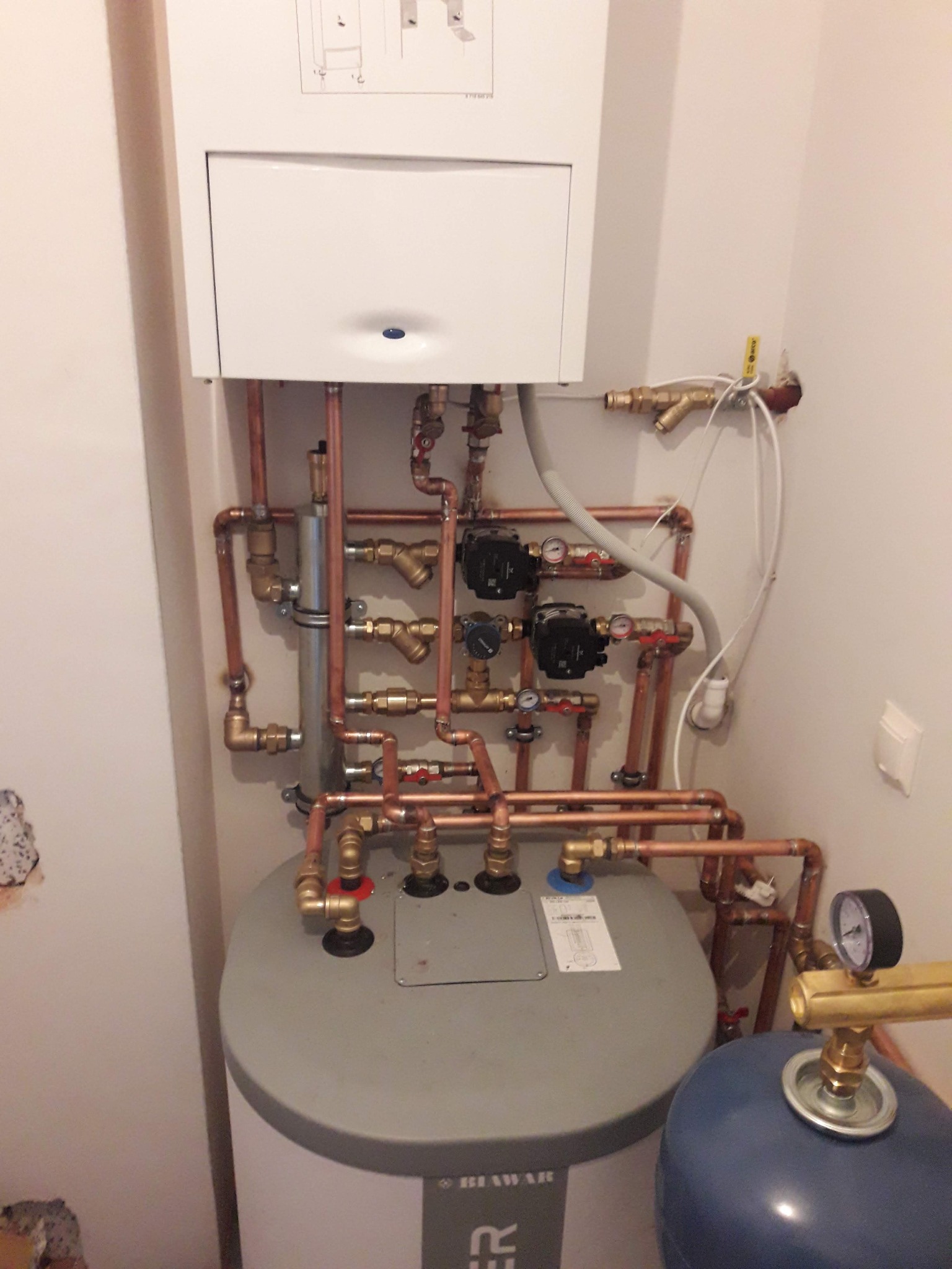 Kocioł grzewczy z rozbudowaną instalacją hydrauliczną z miedzianych rur, zaworów i pomp. Widoczne elementy kotła, zbiornik i osprzęt. Białe ściany w tle.