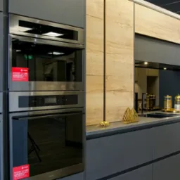 Nowoczesna kuchnia z szarymi szafkami dolnymi i drewnopodobnymi górnymi, z wbudowanymi piekarnikami i dekoracyjnymi akcentami w postaci złotych figurek na blacie.