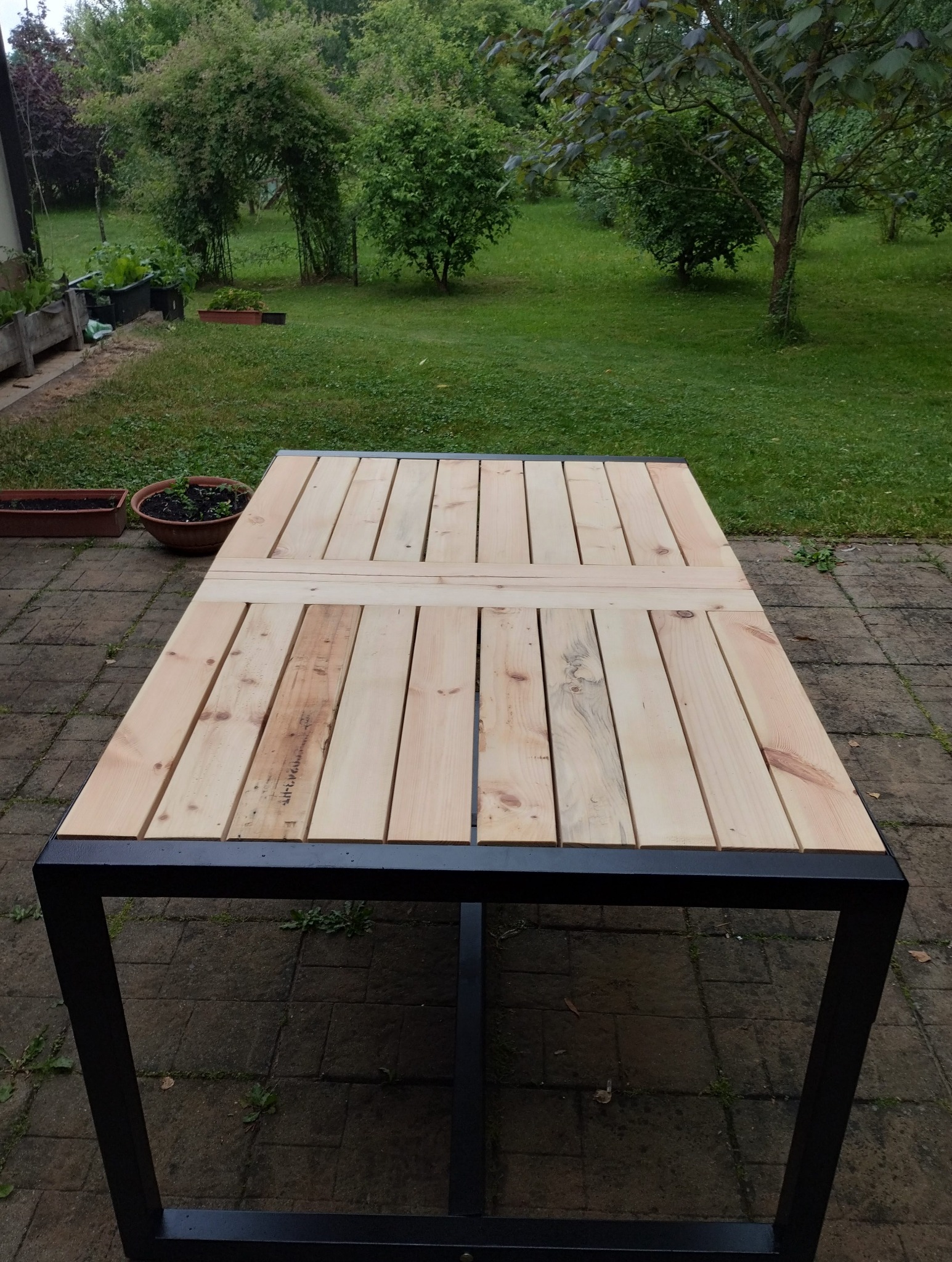 Stół ogrodowy z drewnianym blatem z desek i metalową czarną podstawą, ustawiony na brukowanym patio z widokiem na zielony ogród.