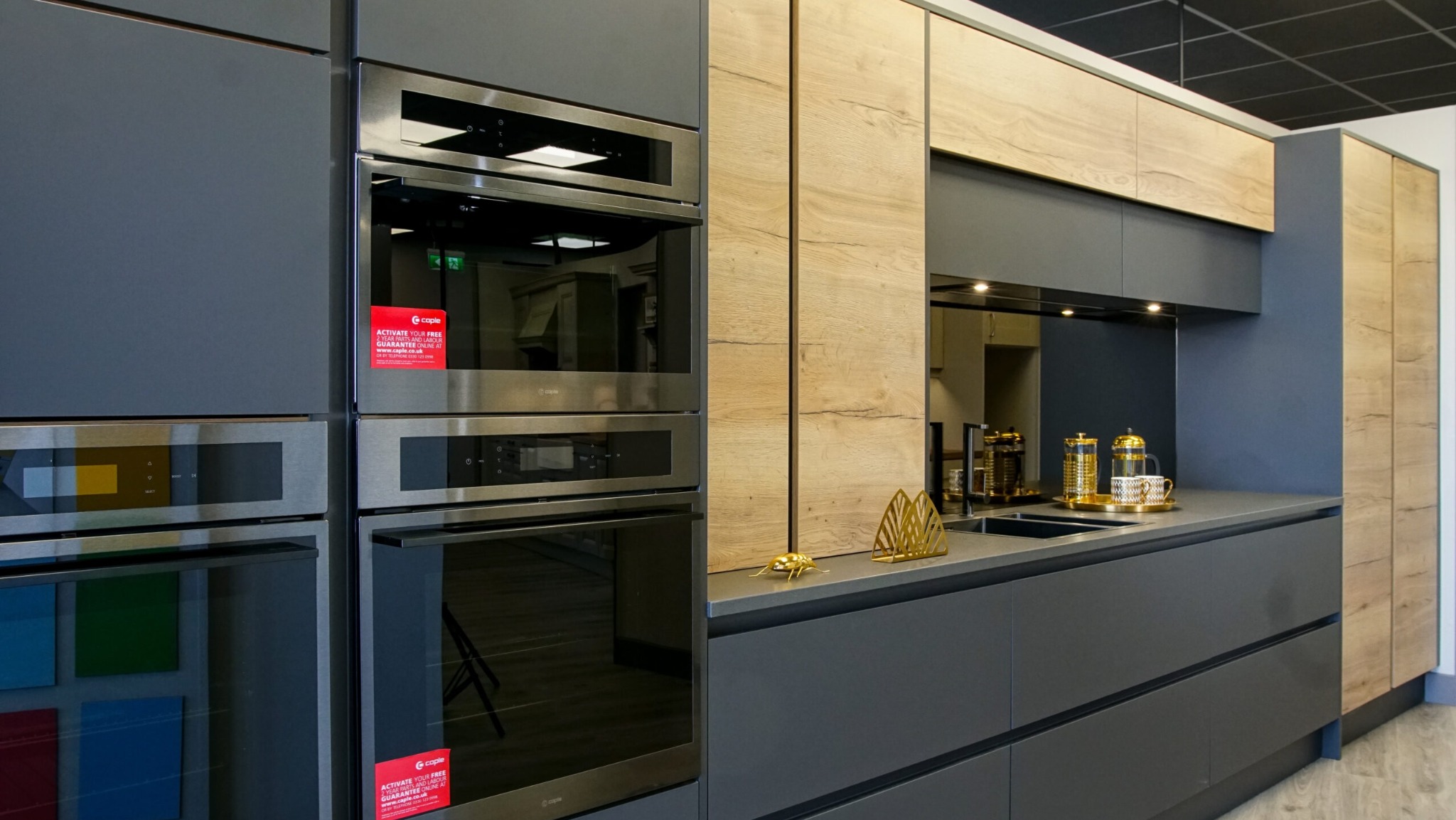 Nowoczesna kuchnia z szarymi szafkami dolnymi i drewnopodobnymi górnymi, z wbudowanymi piekarnikami i dekoracyjnymi akcentami w postaci złotych figurek na blacie.