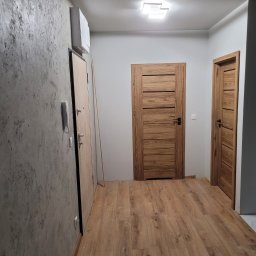 Przemysław Baranowski - Korytarz po remoncie z drewnopodobną podłogą, dwiema drewnianymi drzwiami z poziomymi panelami, szarą ścianą z efektem betonu i białym domofonem.