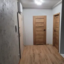 Korytarz po remoncie z drewnopodobną podłogą, dwiema drewnianymi drzwiami z poziomymi panelami, szarą ścianą z efektem betonu i białym domofonem.