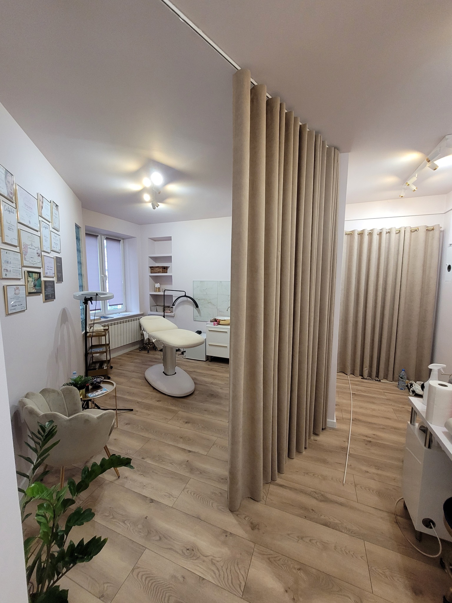 Jasne pomieszczenie z beżowymi zasłonami działowymi, fotelem w kształcie muszli, kozetką kosmetyczną i ścianą z certyfikatami, oświetlone lampami sufitowymi