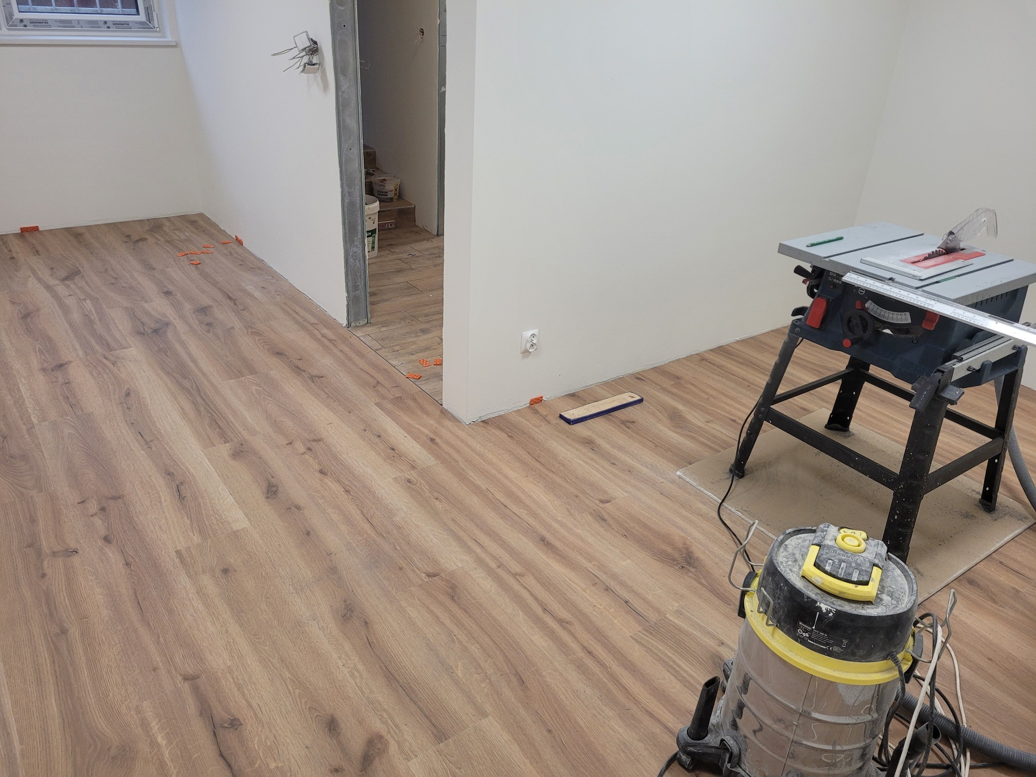 Układanie paneli podłogowych w jasnym pomieszczeniu, widoczna piła stołowa i odkurzacz przemysłowy, pomarańczowe kliny dystansowe wzdłuż ściany.