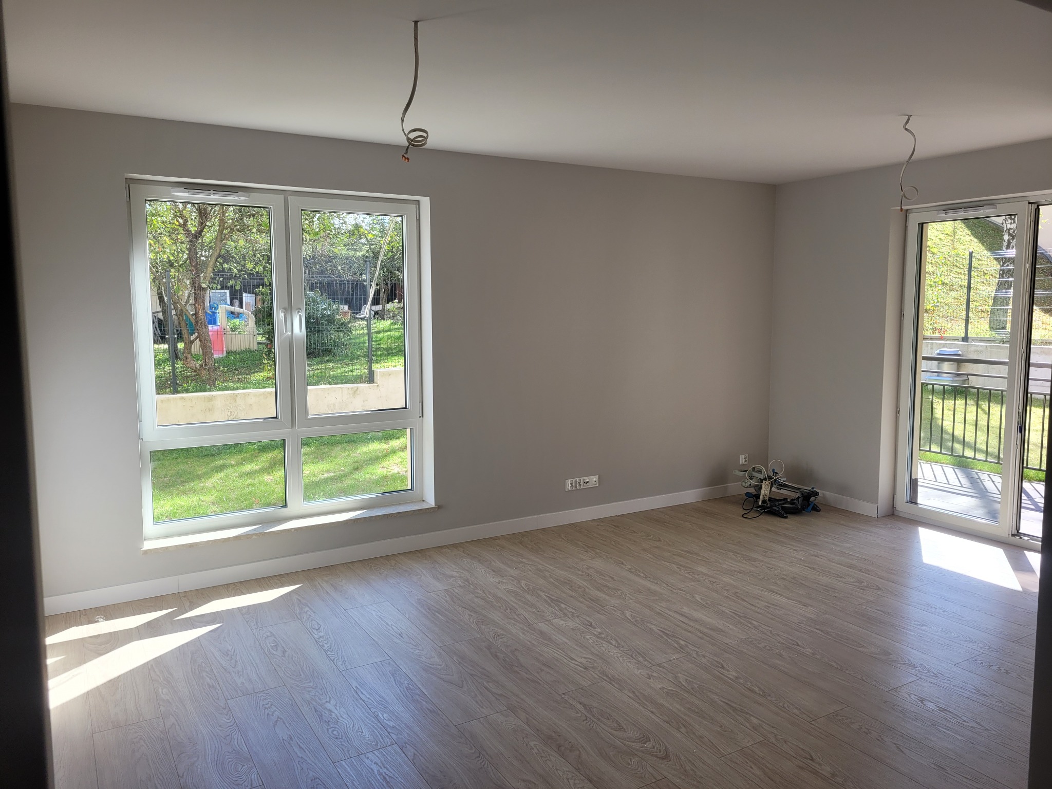 Pusty pokój z panelami podłogowymi, świeżo pomalowanymi szarymi ścianami, dużym oknem z widokiem na ogród i niezakończonymi instalacjami elektrycznymi na suficie, przygotowany do wykończenia.