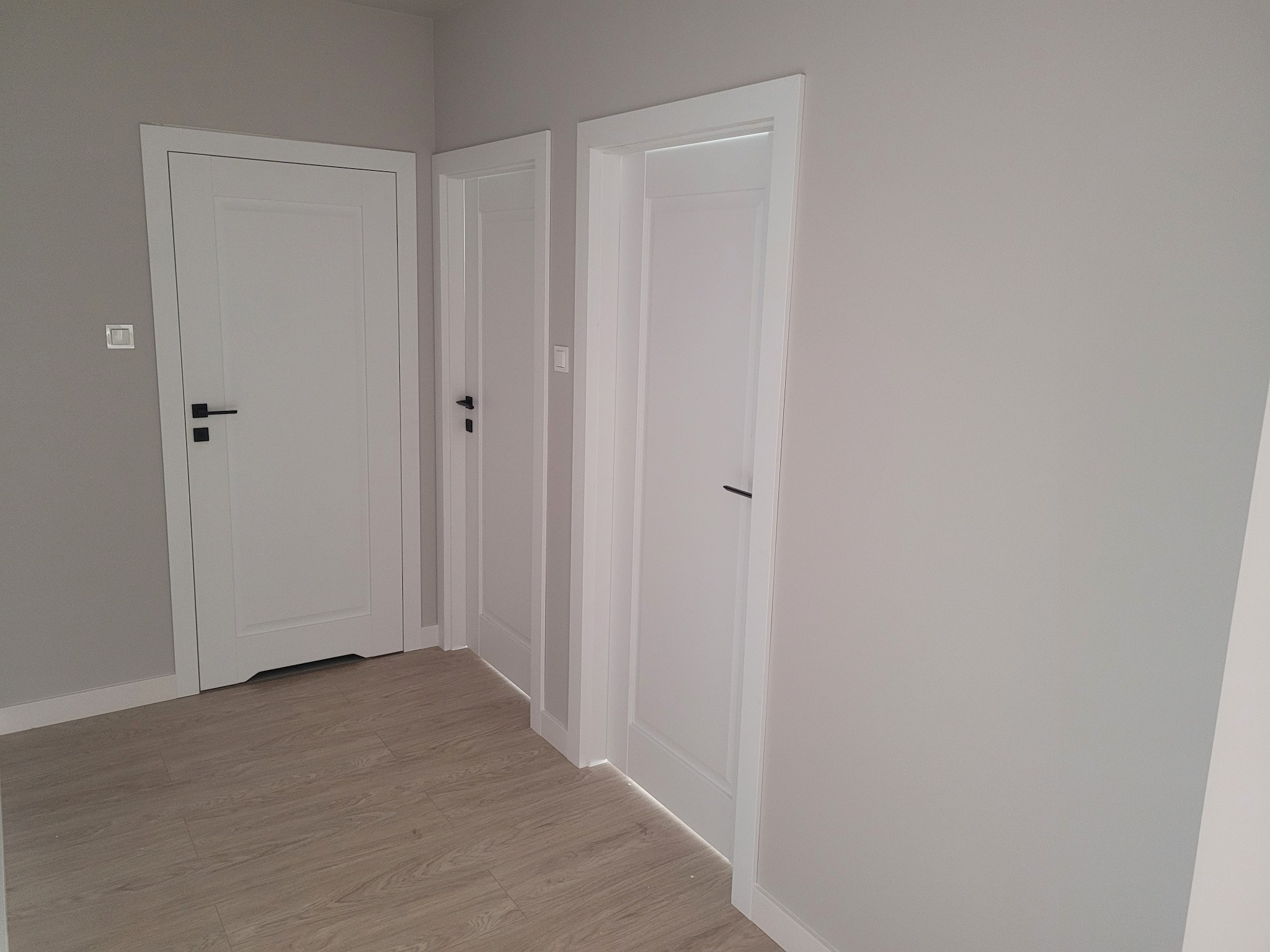 Trzy białe, panelowe drzwi wewnętrzne z czarnymi klamkami na tle szarej ściany i podłogi z paneli w odcieniach beżu, widok z perspektywy.