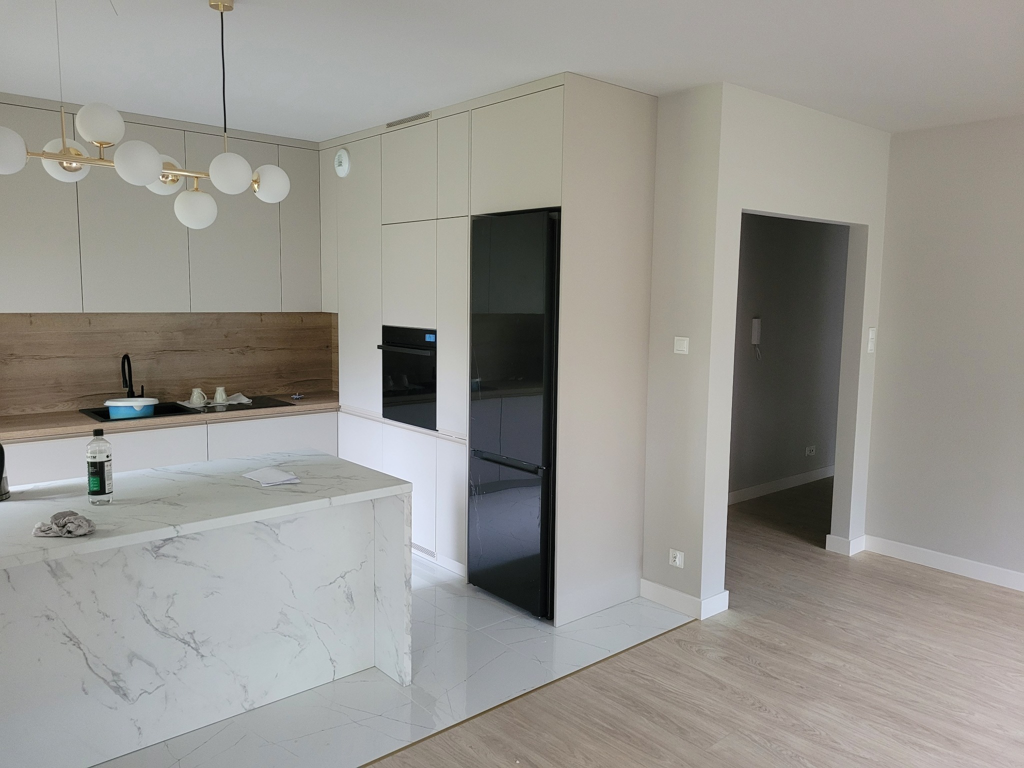 Nowoczesna kuchnia z wyspą wykończoną marmurem, zabudową w odcieniach szarości i czarną lodówką, oświetlona lampą z kulistymi kloszami; widoczne akcesoria i środki czystości na blacie.