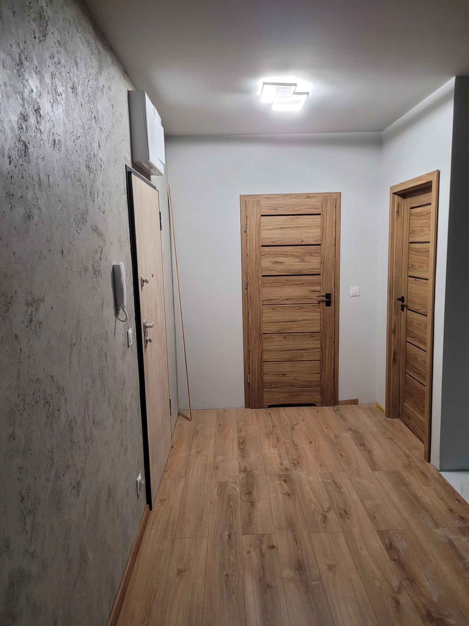 Korytarz po remoncie z drewnopodobną podłogą, dwiema drewnianymi drzwiami z poziomymi panelami, szarą ścianą z efektem betonu i białym domofonem.