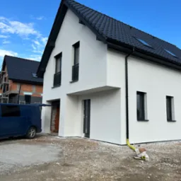 Nowoczesny, biały dom z czarnym dachem i oknami. Budynek częściowo wykończony, z widocznym fragmentem elewacji w trakcie prac. Na podjeździe stoi niebieski bus.
