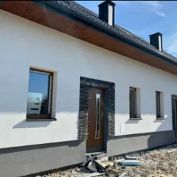 Elewacja domu jednorodzinnego z oknami w brązowych ramach i drzwiami wejściowymi obłożonymi kamieniem dekoracyjnym. Widoczne wykończenie cokołu w szarym kolorze.