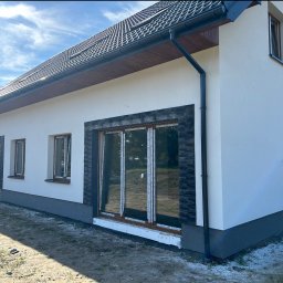 F.r.b.domino - Elewacja domu z oknami w brązowych ramach, wykończona kamieniem dekoracyjnym i pomalowana na biało. Widoczne orynnowanie i fragment dachu z dachówką.