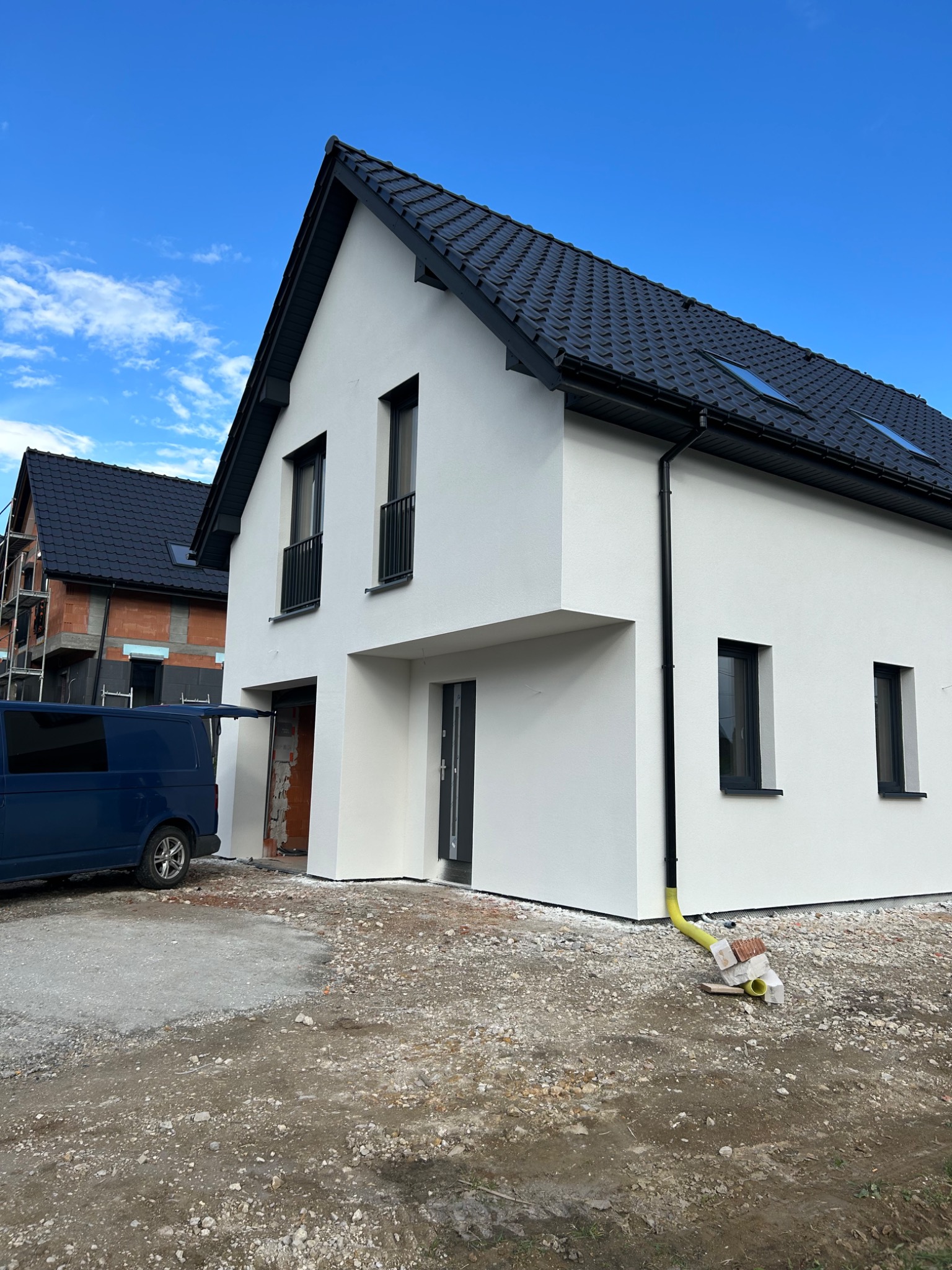 Nowoczesny, biały dom z czarnym dachem i oknami. Budynek częściowo wykończony, z widocznym fragmentem elewacji w trakcie prac. Na podjeździe stoi niebieski bus.