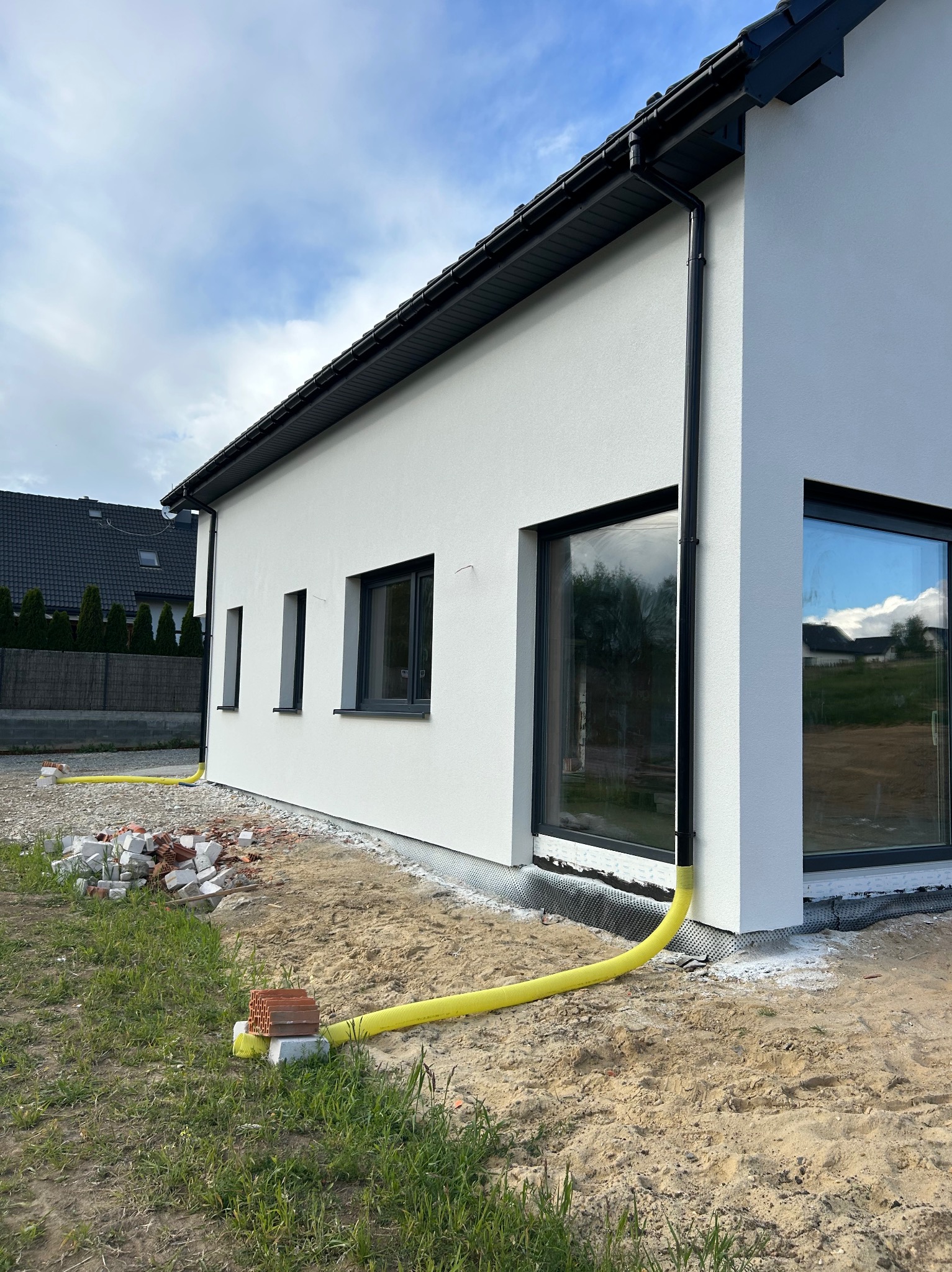 Nowoczesny dom jednorodzinny z białym tynkiem i grafitową stolarką okienną, widok z zewnątrz, z żółtą rurą drenażową i stertą cegieł w tle.