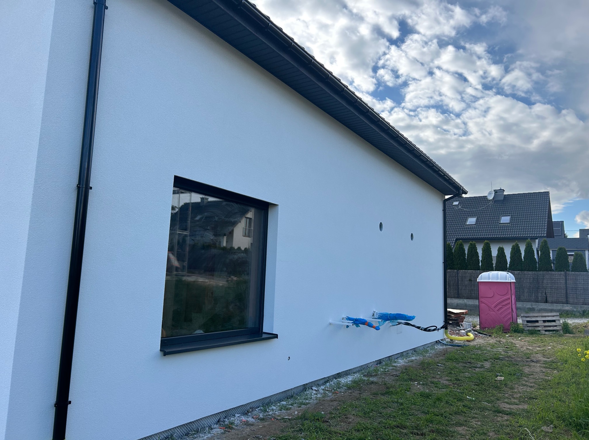 Biała elewacja domu jednorodzinnego z czarną stolarką okienną i rurą spustową. Widoczne wystające rury zabezpieczone folią. W tle inne domy i niebo z chmurami.