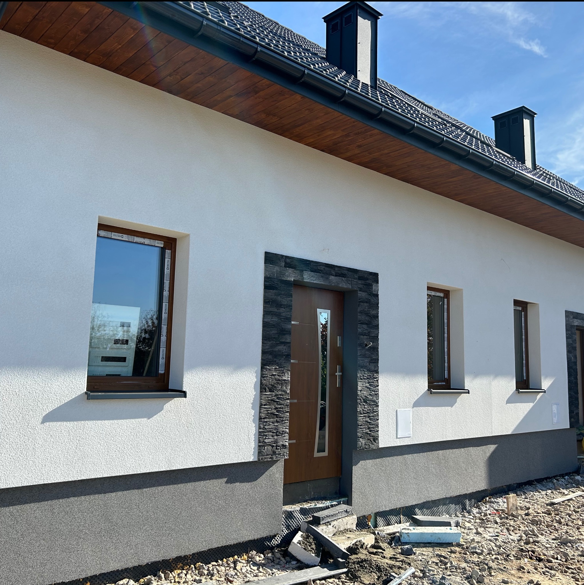 Elewacja domu jednorodzinnego z oknami w brązowych ramach i drzwiami wejściowymi obłożonymi kamieniem dekoracyjnym. Widoczne wykończenie cokołu w szarym kolorze.