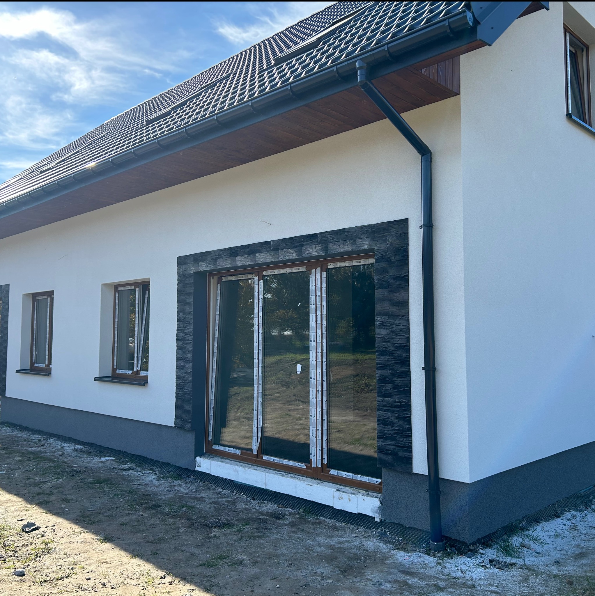 Elewacja domu z oknami w brązowych ramach, wykończona kamieniem dekoracyjnym i pomalowana na biało. Widoczne orynnowanie i fragment dachu z dachówką.