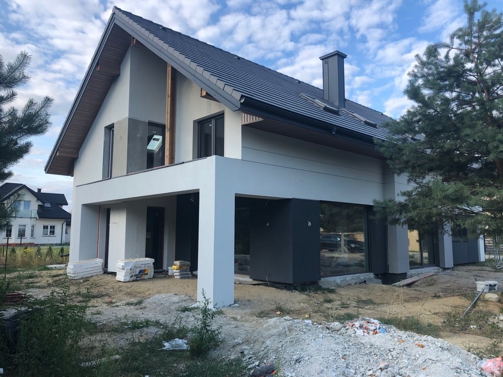 Nowoczesny dom jednorodzinny z szarą elewacją i ciemną dachówką, widoczne materiały budowlane na posesji, sugerujące prace wykończeniowe.