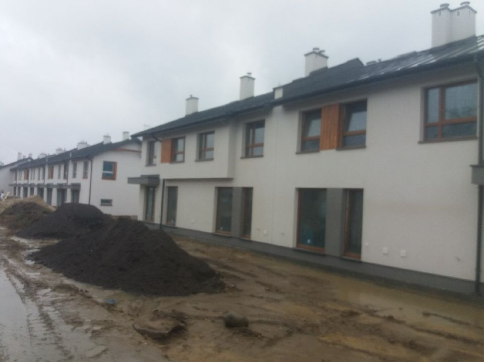 Rząd nowych, dwukondygnacyjnych domów szeregowych z białymi elewacjami i ciemnymi dachami, widoczny w pochmurny dzień. Na pierwszym planie hałdy ziemi i błotniste podłoże.