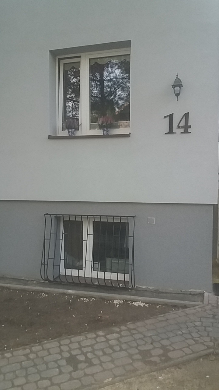 Dwukondygnacyjny fragment elewacji budynku z oknami, numerem '14' i lampą. Okno piwniczne z kratą, nad nim okno z kwiatami. Jasna elewacja, szara podmurówka.